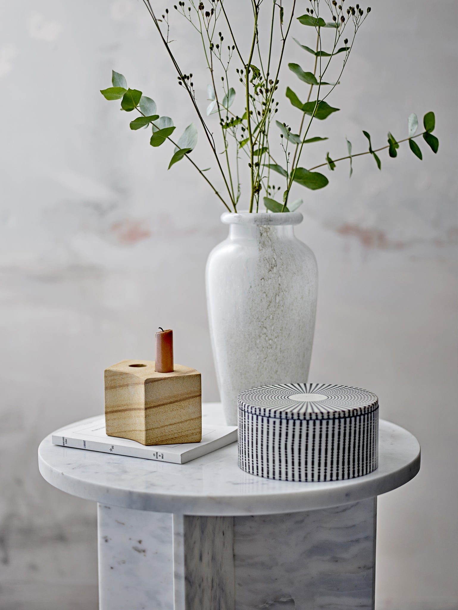 Un tavolino tondo Jasmia in marmo bianco di Divani.Store presenta un vaso in ceramica bianca pieno di rami lunghi e sottili e foglie verdi. Lo accompagna un portacandele in legno con una candela rossa e una scatola rotonda con motivi, il tutto accentuando l'arredamento di design nordico contro la parete chiara e strutturata.