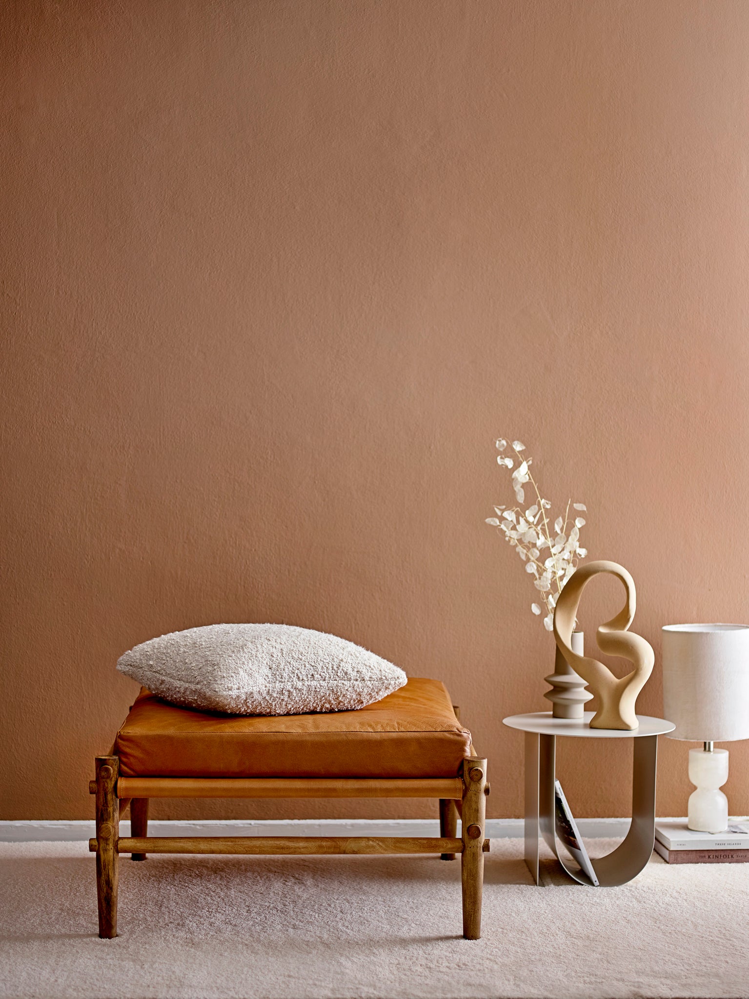 Un interno minimalista mette in mostra una panca di legno con un cuscino marrone e un soffice cuscino bianco. Accanto c'è il tavolino scandinavo Cher in metallo con spazio portariviste grigio chiaro di Divani.Store, che espone sculture astratte e fiori secchi. Nelle vicinanze c'è una lampada color crema, il tutto su uno sfondo di una parete calda e marrone e un pavimento in moquette chiara.