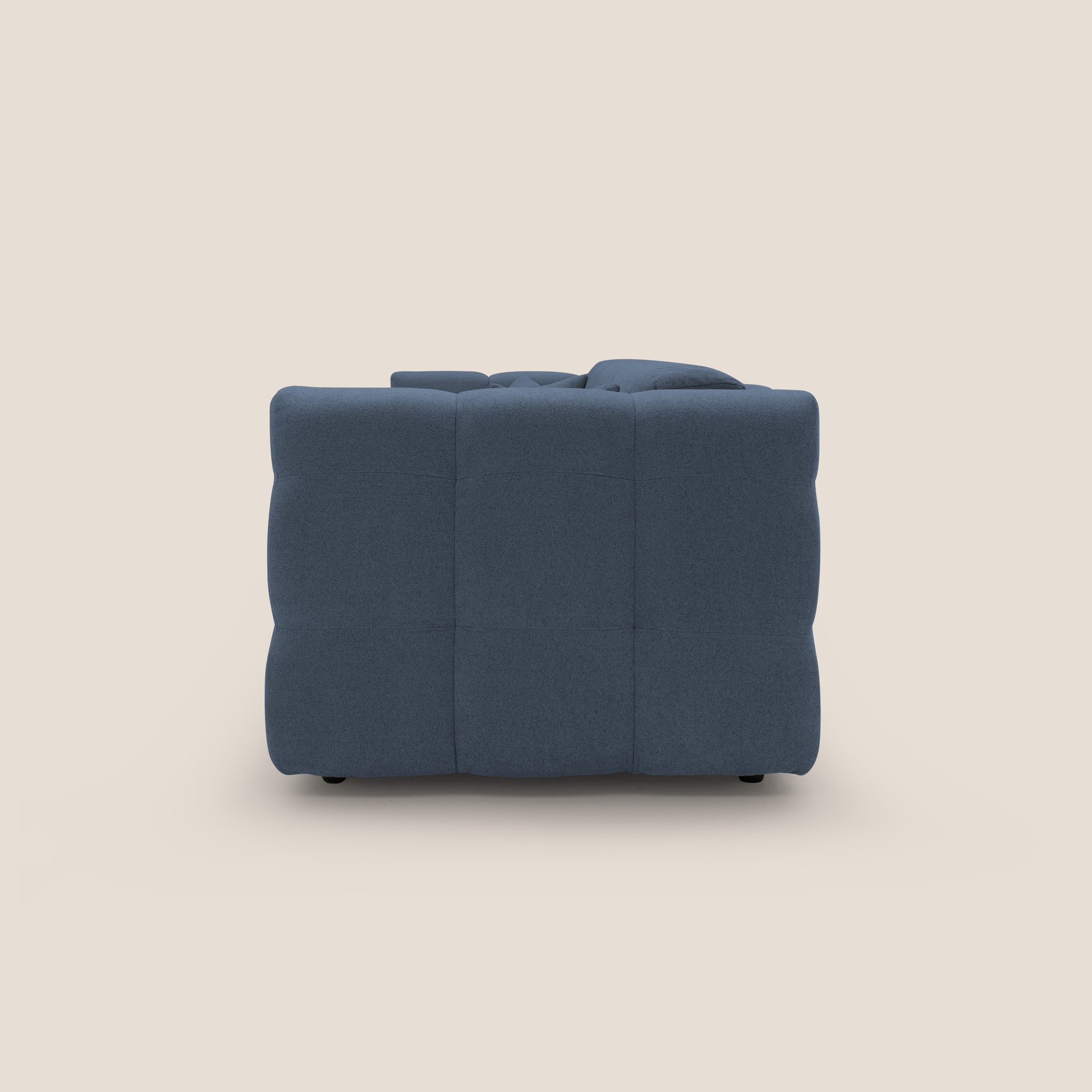 La poltrona Tender di Divani.Store è esposta di lato su uno sfondo beige, che mette in mostra il suo soffice tessuto blu scuro, il design capitonné e la tappezzeria imbottita. Unisce il comfort allo stile italiano moderno, enfatizzando la sua lussuosa consistenza e il suo stile contemporaneo.