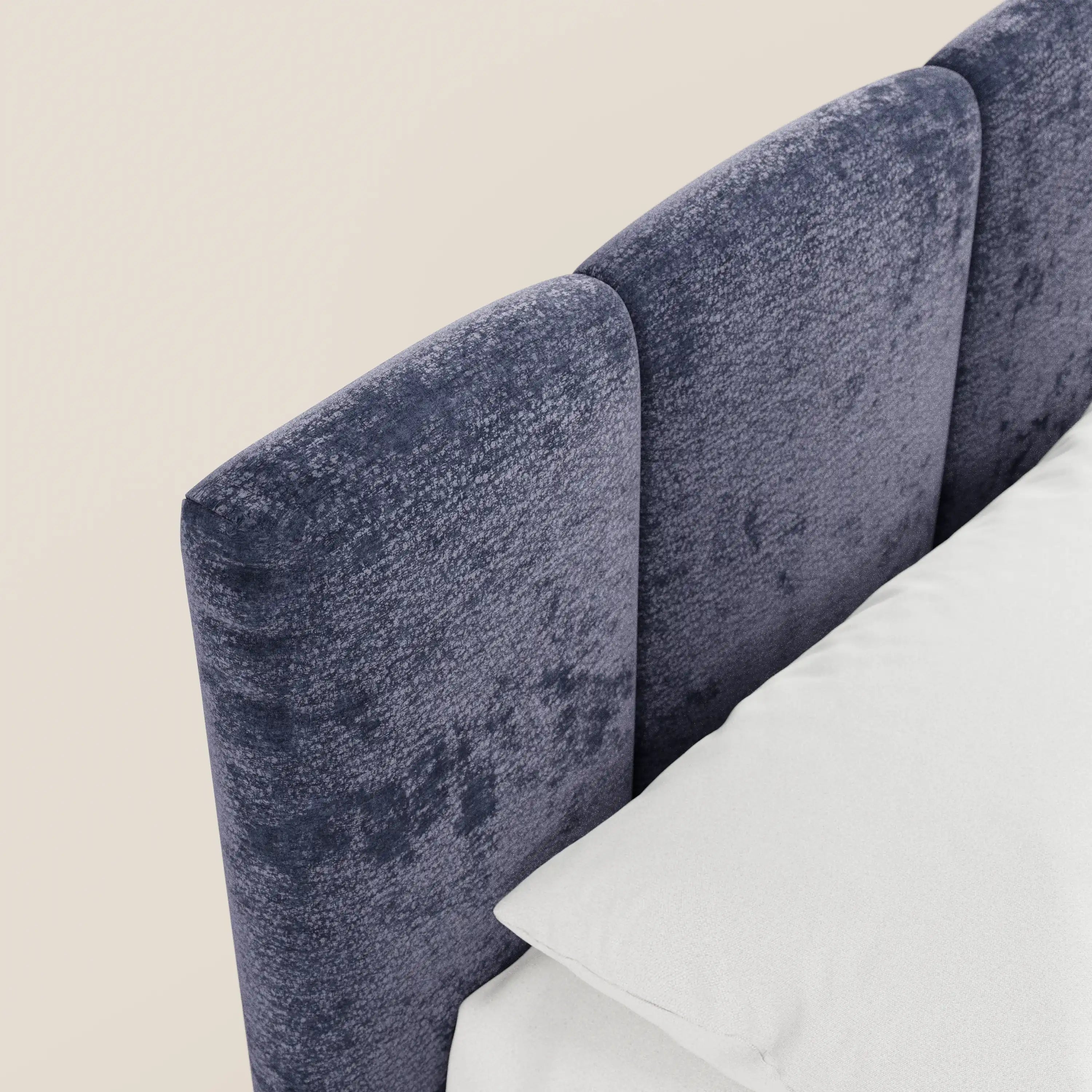 Un primo piano del letto imbottito ARKAM di Letti.store mette in risalto la testiera con cuciture verticali e texture, realizzata in tessuto blu scuro certificato Oeko-Tex®. Sulla destra si intravedono un cuscino bianco e parte della biancheria da letto, che si stagliano su uno sfondo beige chiaro, creando un effetto shabby chic.