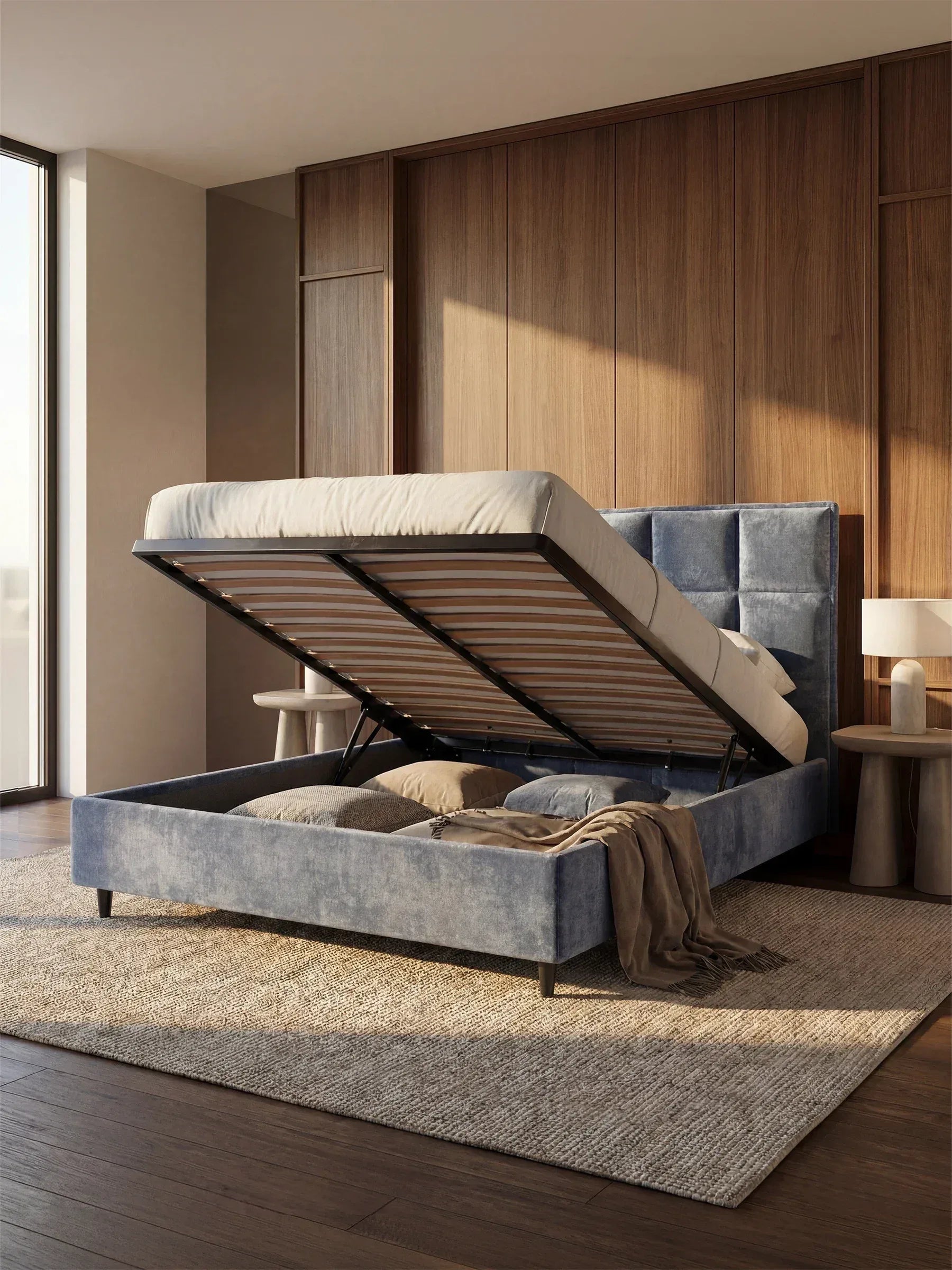 Una camera da letto moderna caratterizzata da un'ampia finestra, pareti rivestite in legno e dal letto imbottito AURO con contenitore e testiera capitonné alta in ciniglia Oeko-Tex® di Letti.store, raffigurato con la base imbottita aperta per mostrare l'ampio spazio di stivaggio sotto il letto. La biancheria da letto dai toni neutri e un morbido plaid completano il letto, mentre due comodini rotondi e un tappeto beige completano l’arredamento accogliente.