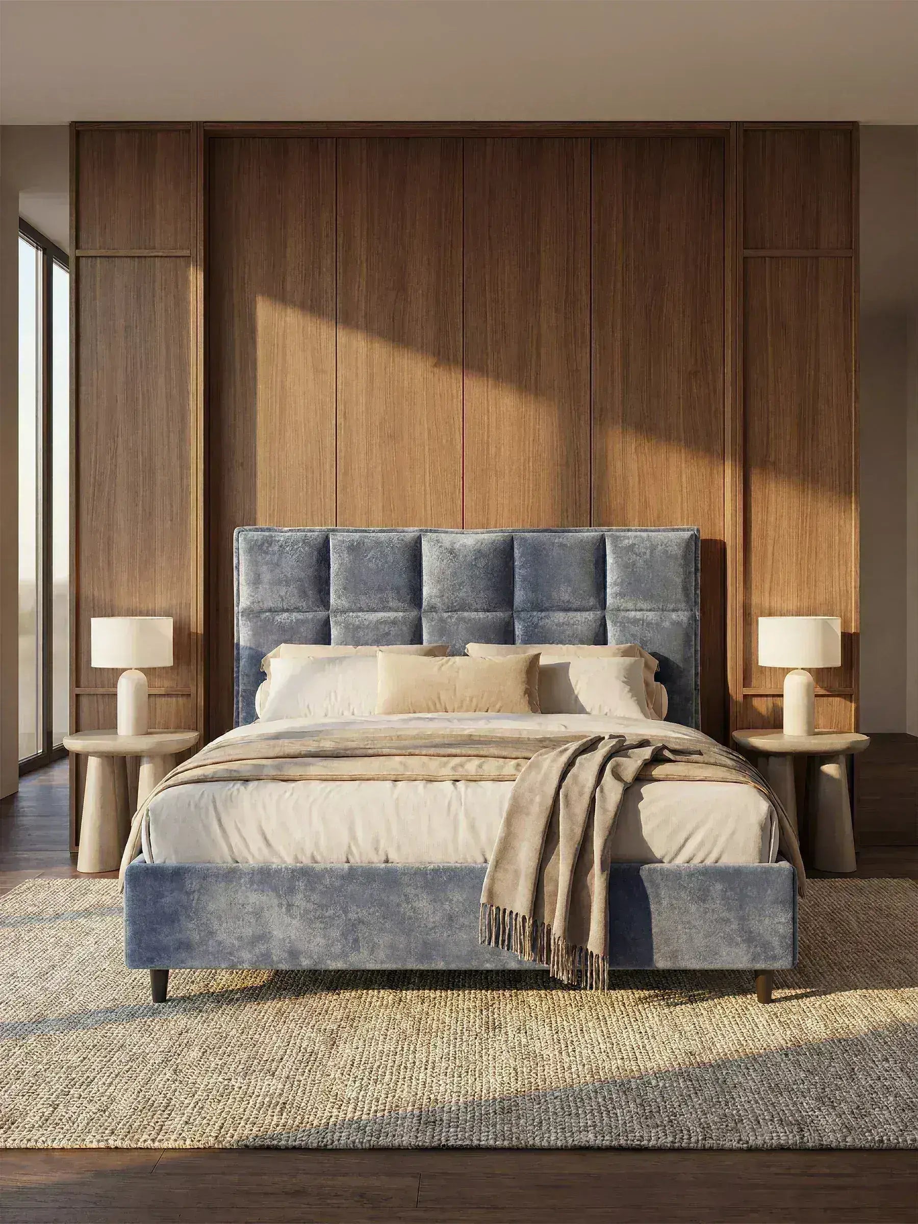 Una camera da letto moderna arredata con il letto imbottito AURO con contenitore e testiera capitonné alta in ciniglia Oeko-Tex® di Letti.store, in velluto blu, abbinato a biancheria da letto beige, due lampade beige sui comodini in legno e un tappeto dalla trama rigata. La parete dietro al letto è impreziosita da un rivestimento verticale in legno, mentre la luce del sole filtra da una finestra sulla sinistra.