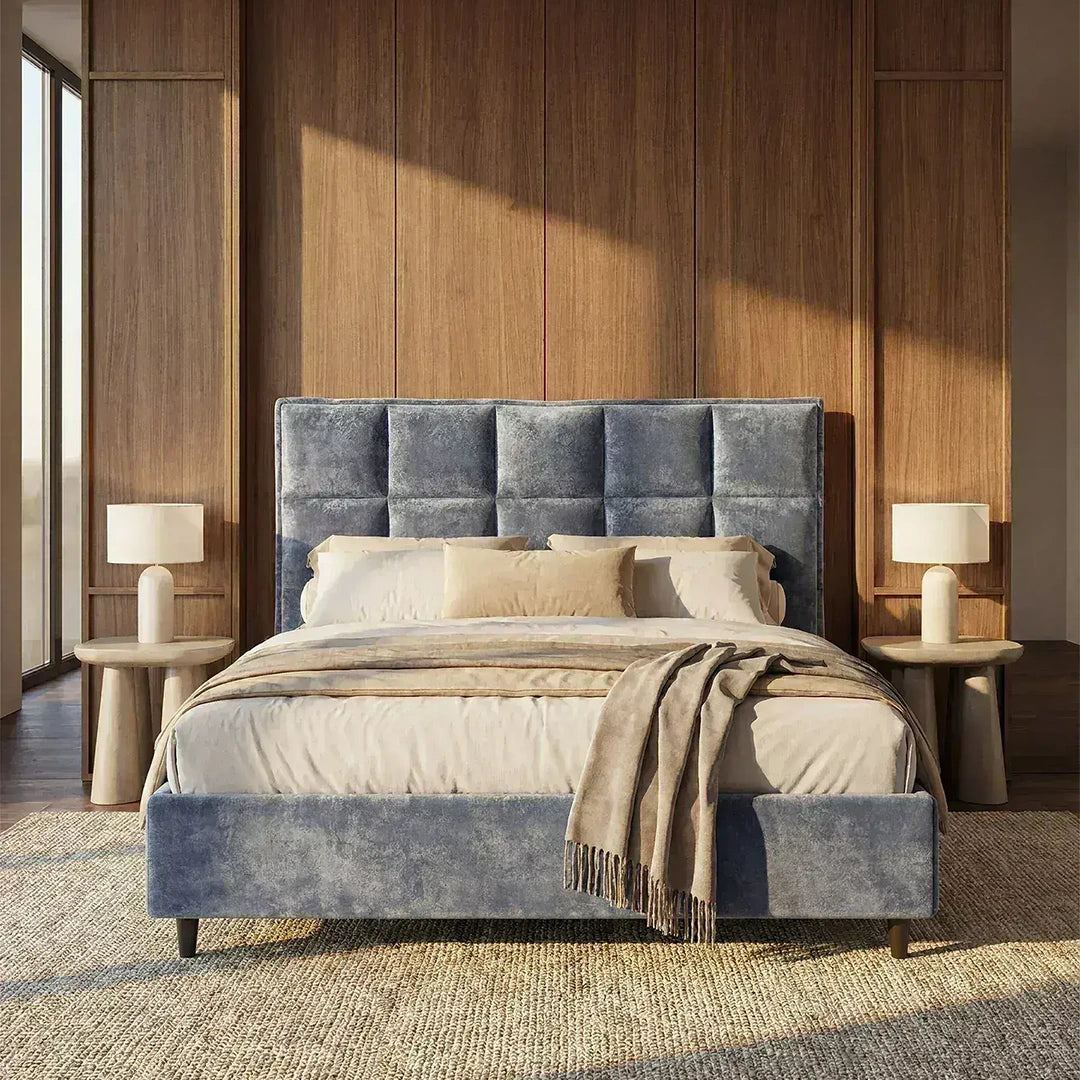 Una camera da letto moderna con il letto imbottito AURO con contenitore e testiera capitonné alta in ciniglia Oeko-Tex® di Letti.store, biancheria da letto beige, un copriletto color cuoio, due comodini rotondi in legno con lampade bianche, rivestimento verticale in legno e luce naturale.