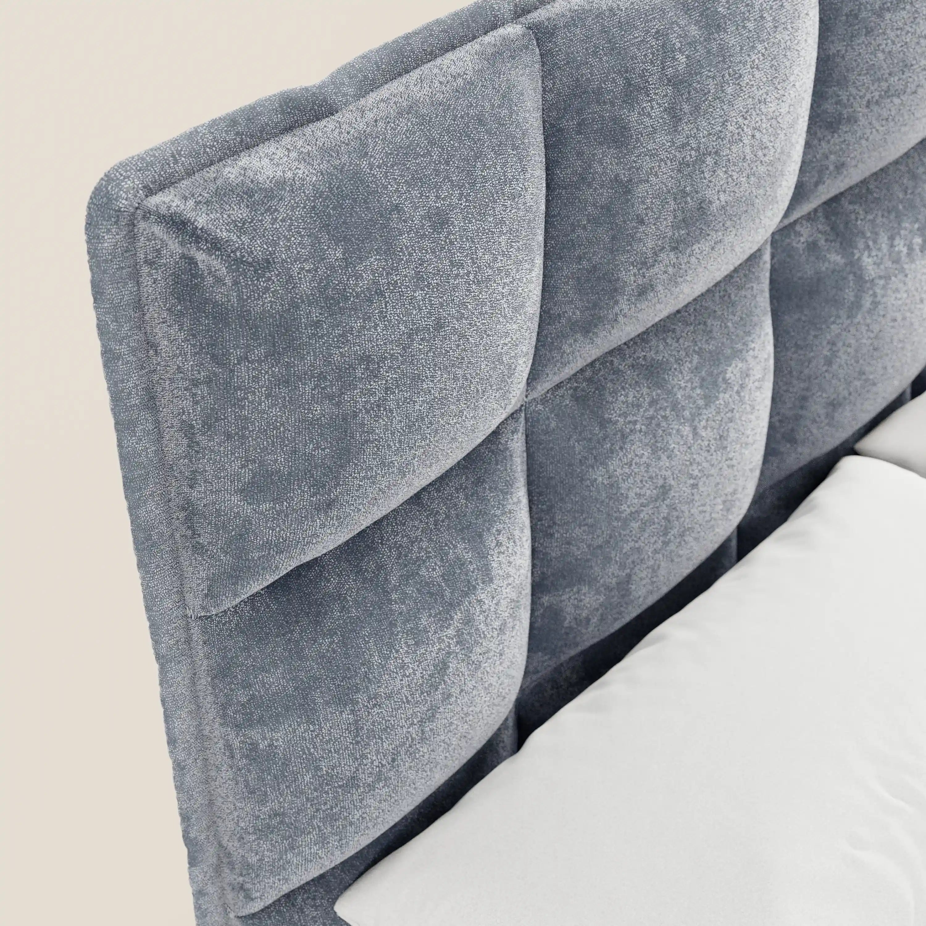 Primo piano del letto imbottito AURO di Letti.store, caratterizzato da un’alta testiera capitonné in morbida ciniglia grigia certificata Oeko-Tex®. L’immagine mostra l’angolo della testiera e un cuscino bianco su uno sfondo beige chiaro.