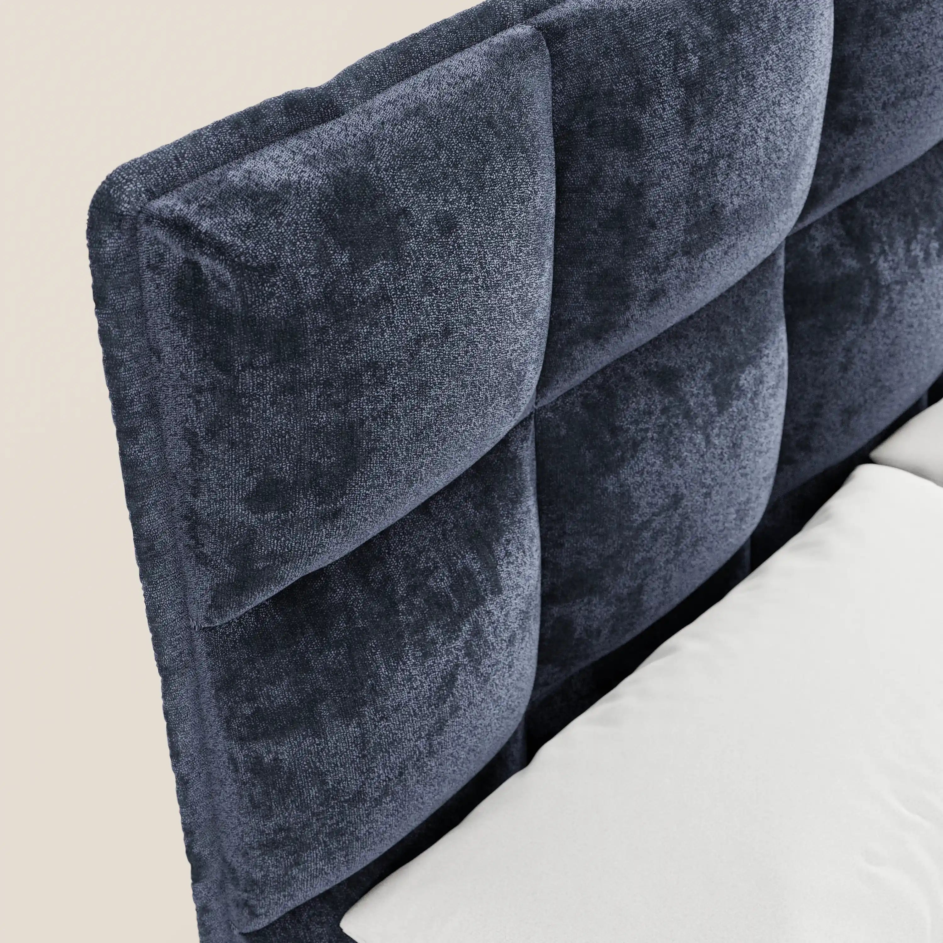 Primo piano del letto imbottito AURO di Letti.store, caratterizzato da una testiera capitonné in ciniglia blu scuro certificata Oeko-Tex® con morbidi quadrati imbottiti. Un cuscino bianco è appoggiato contro l'alta testiera; lo sfondo è di un colore chiaro a tinta unita.