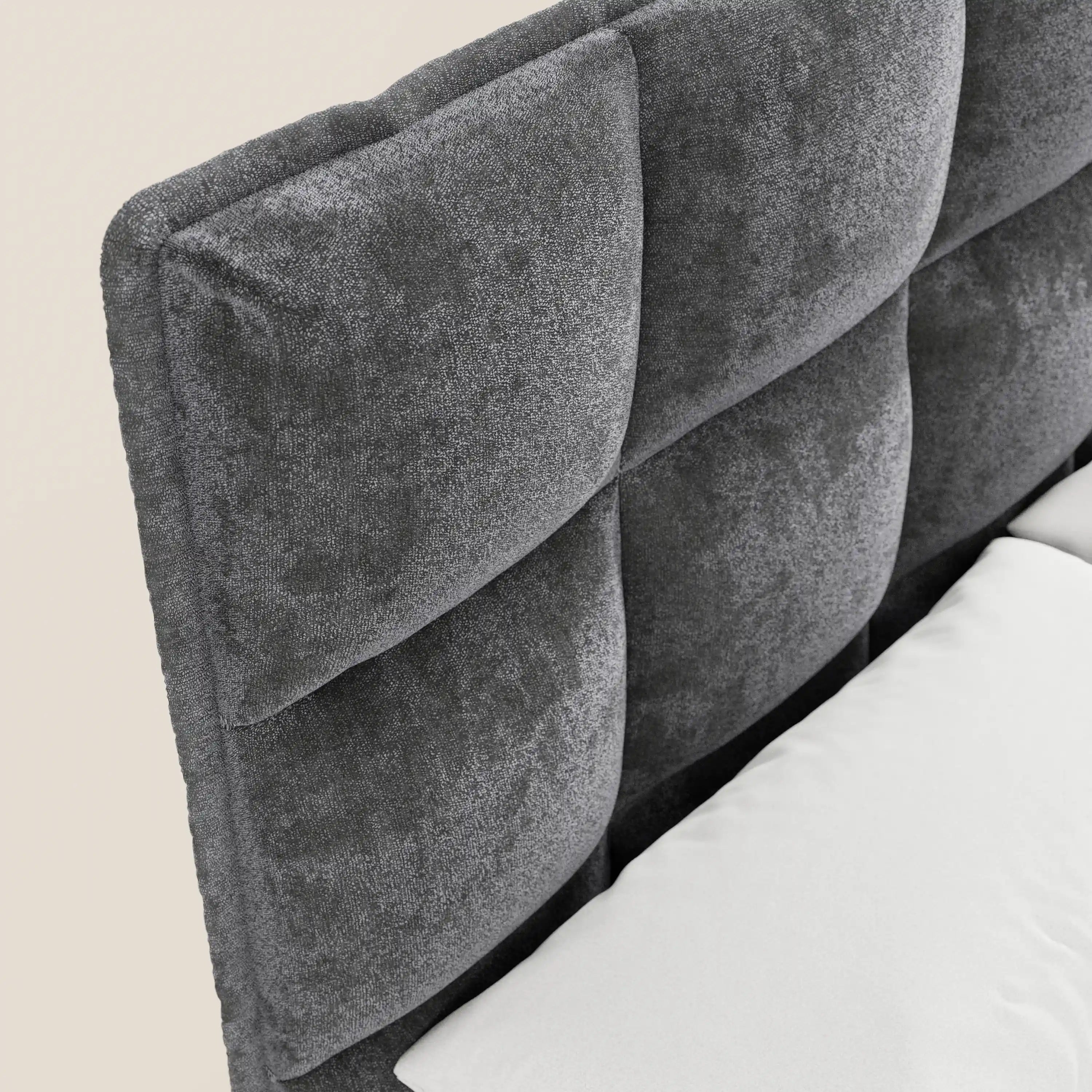 Un primo piano del letto imbottito AURO di Letti.store mette in risalto la sua testiera alta capitonné in ciniglia Oeko-Tex®, rivestita in morbido tessuto grigio scuro con texture e dotata di ampi pannelli imbottiti. In basso a destra, su uno sfondo neutro, spicca un cuscino bianco.