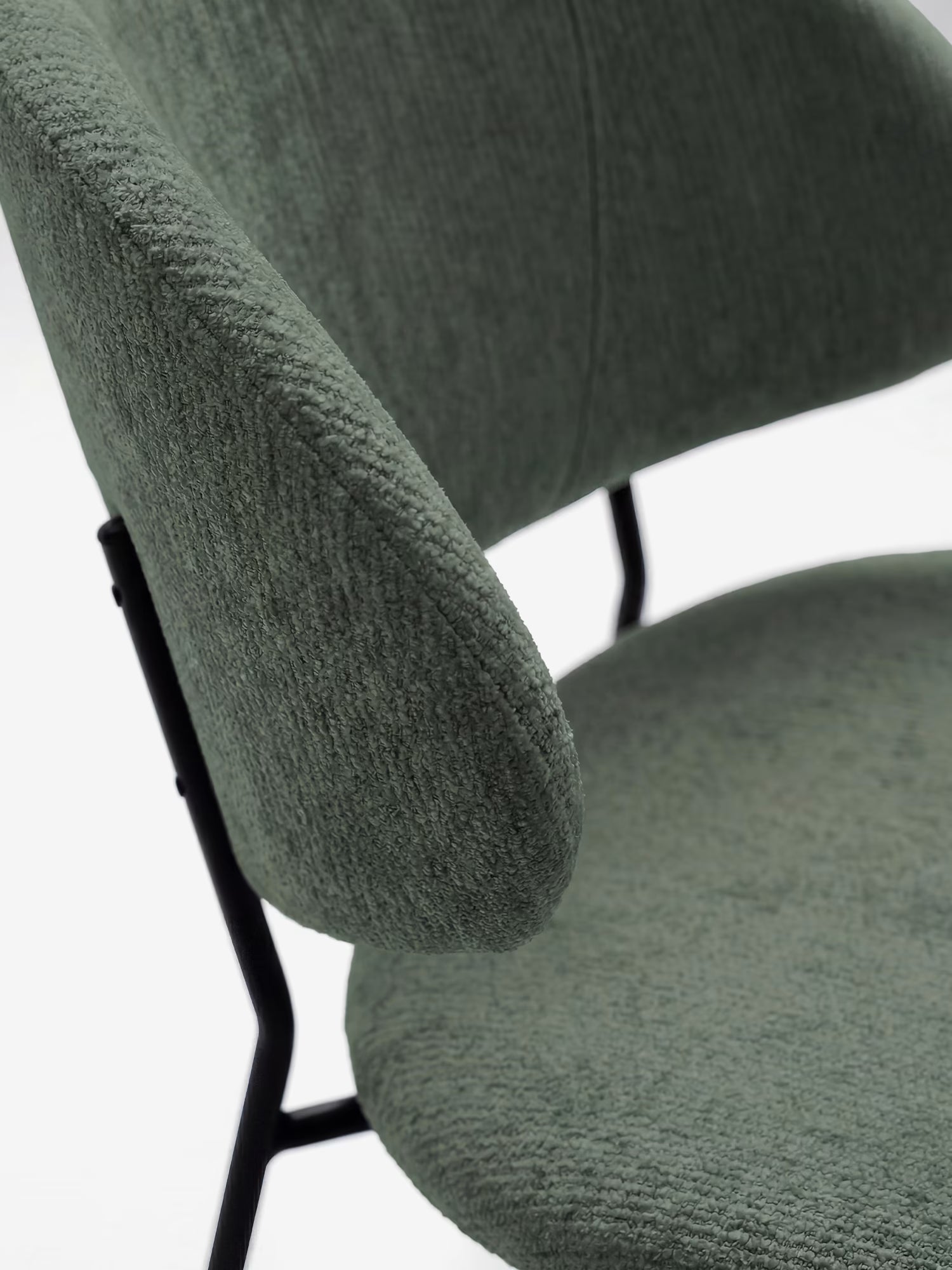 Primo piano della sedia Bruma di Divani.Store: design minimalista con rivestimento verde salvia a trama ruvida, schienale imbottito avvolgente e robuste gambe in metallo nero. Lo sfondo semplice ne esalta il fascino contemporaneo.