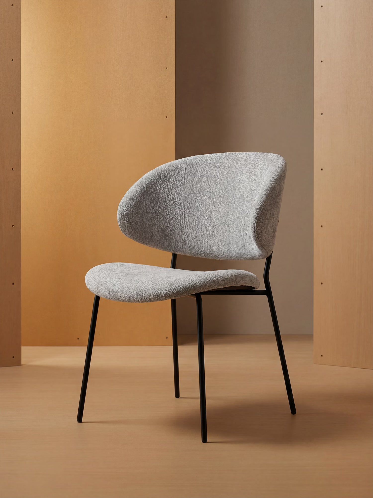 La Bruma di Divani.Store è una sedia realizzata in elegante tessuto goffrato grigio chiaro, con schienale avvolgente e gambe sottili in metallo nero, perfetta per ambienti minimalisti con pannelli e pavimenti effetto legno beige o tan.