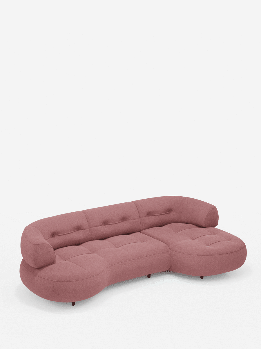 Un divano angolare rosa a forma di fagiolo, modello Croissant, con schienali scorrevoli in morbido tessuto effetto cotone smacchiabile T33, caratterizzato da una superficie trapuntata dalla consistenza morbida, una forma modulare sinuosa e schienali bassi su piccole gambe scure.