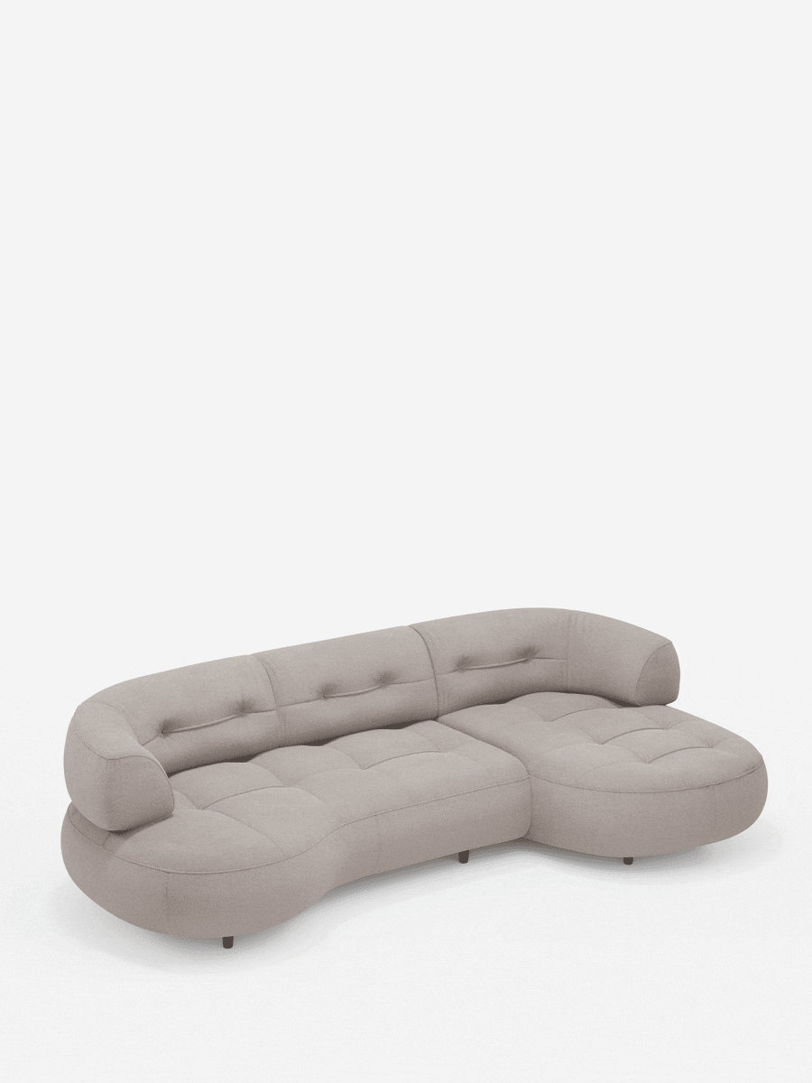 Un divano angolare Croissant beige, dal design a forma di fagiolo, con schienali scorrevoli in morbido tessuto effetto cotone smacchiabile T33, dai bordi arrotondati, cuscini trapuntati e schienale curvo, su piedini bassi e scuri, su uno sfondo bianco.