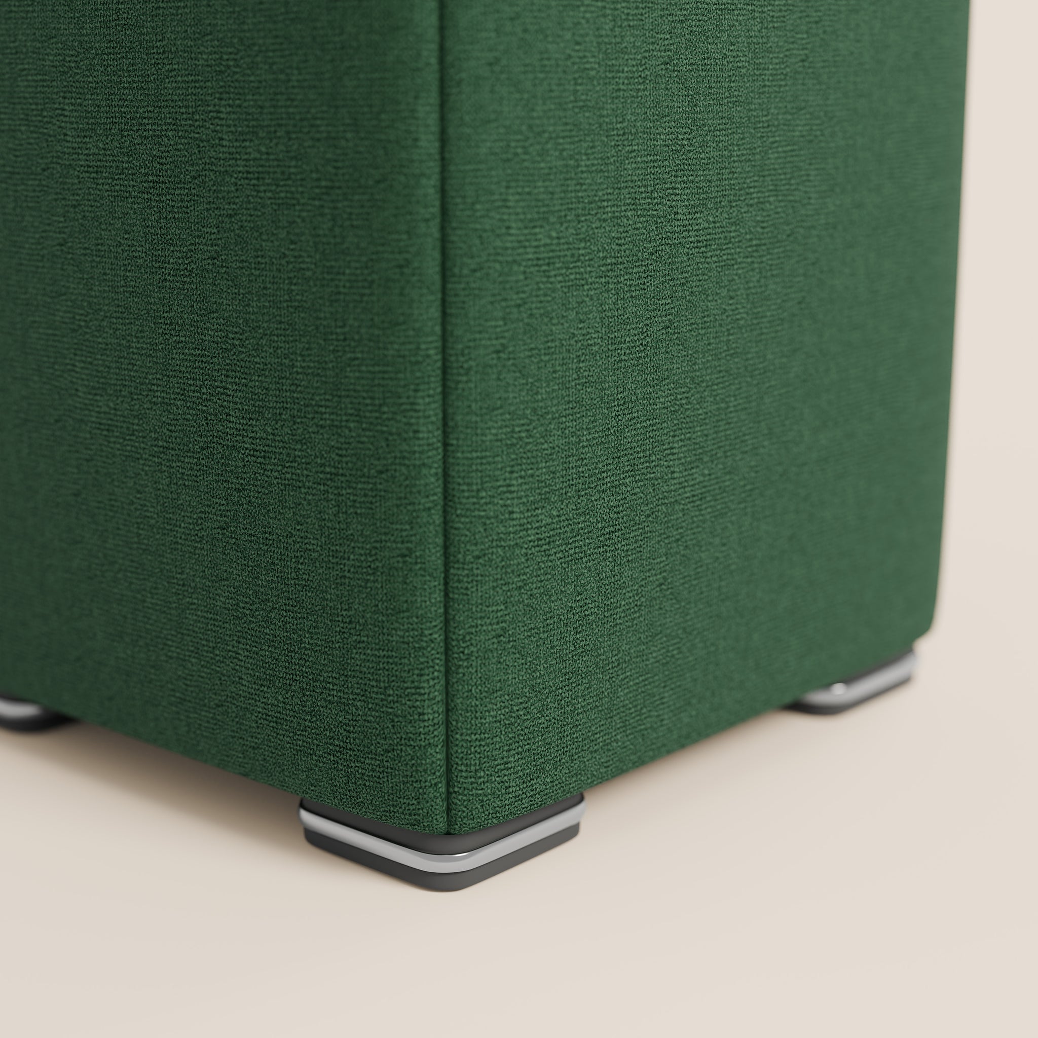 Primo piano di un angolo rivestito in tessuto verde del pouf Cube in tessuto morbido impermeabile T08 VERDE di Divani.Store, che mette in risalto il tessuto strutturato e gli eleganti piedini argentati e neri su una superficie beige chiaro, evidenziando elementi di design moderno.