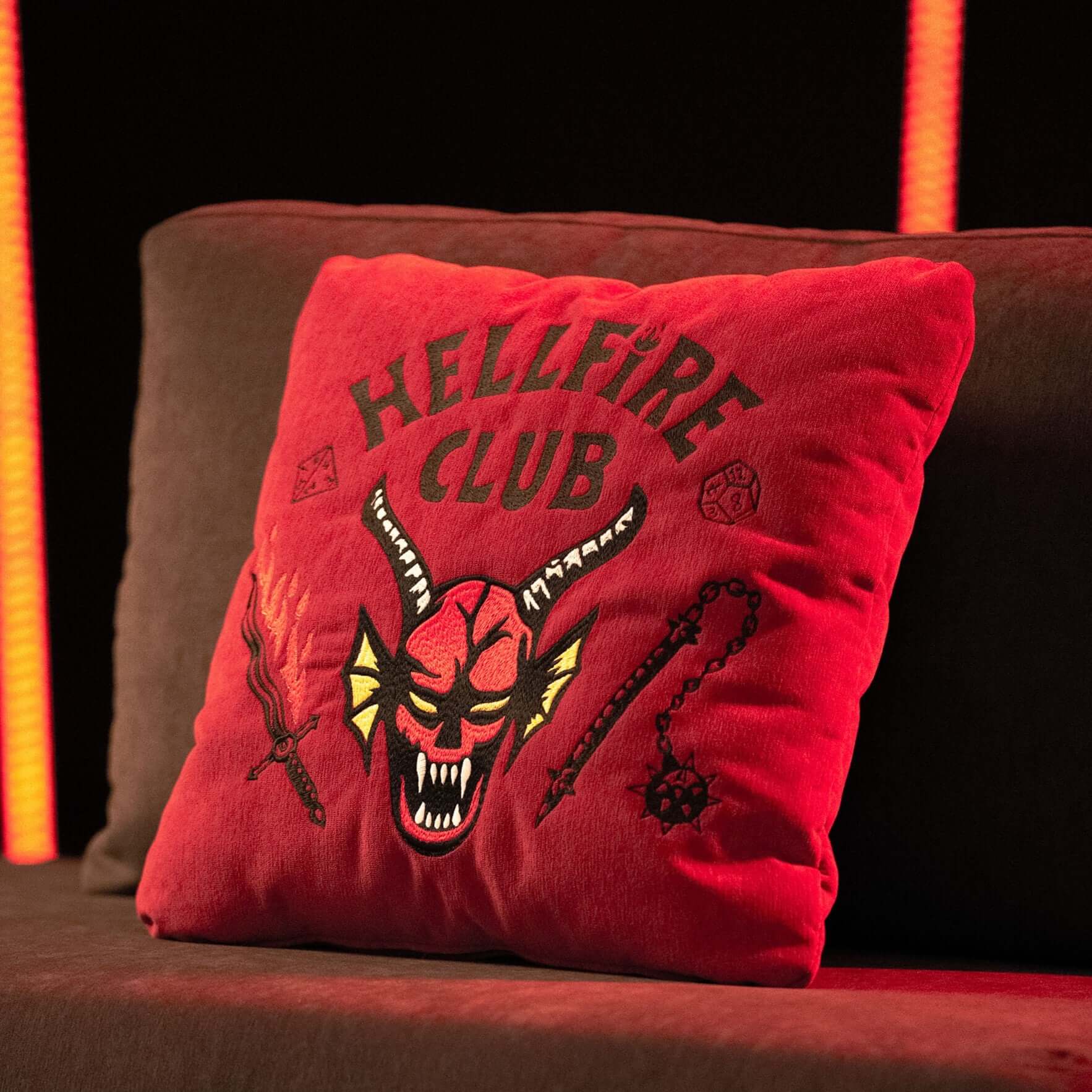 Il cuscino Hellfire Club di Divani.Store, in edizione limitata 45x45 cm, con ricamo "HELLFIRE CLUB", volto demoniaco e motivi fantasy, si illumina sotto le luci rosse verticali, perfetto per i fan di Stranger Things.