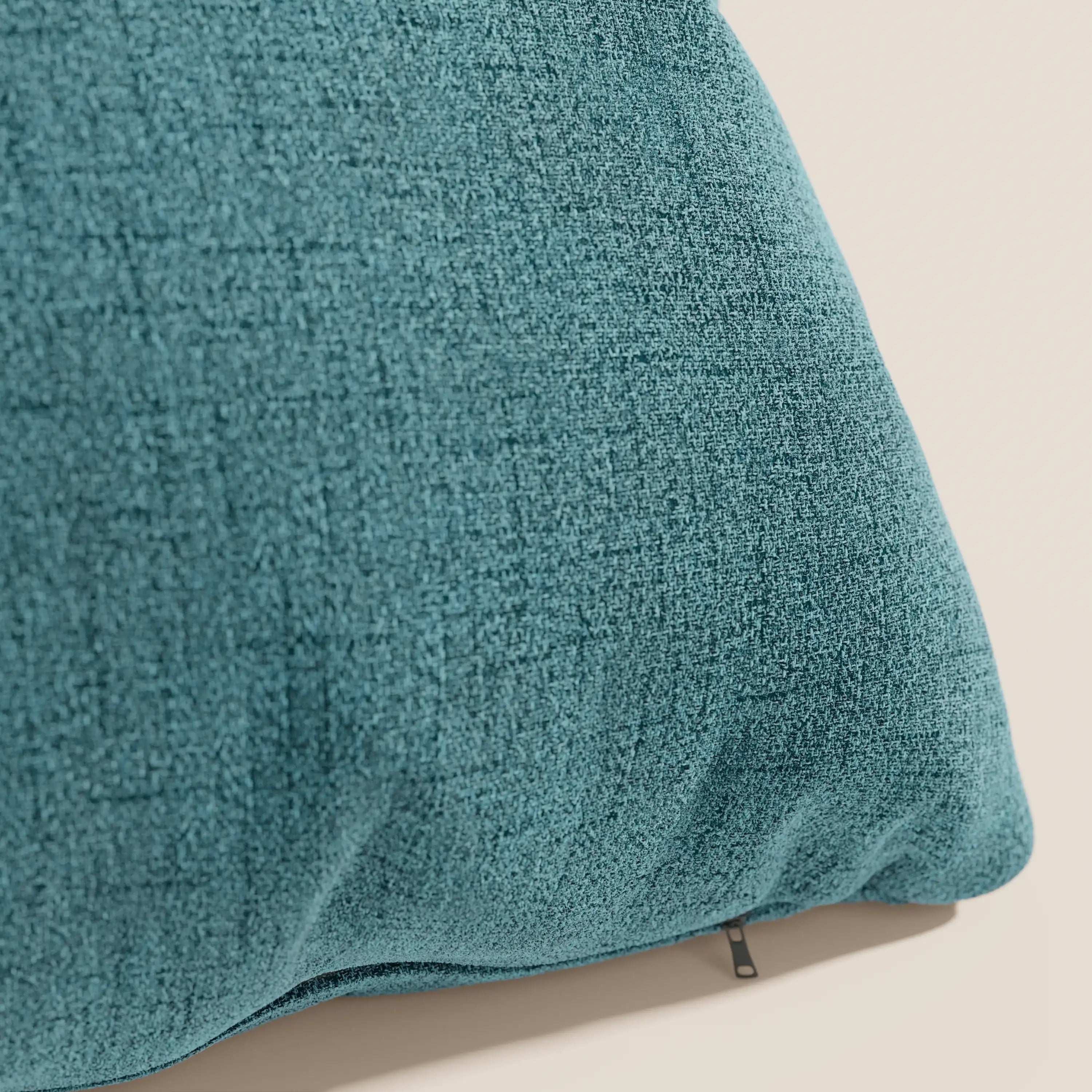 Primo piano del cuscino Felis di Divani.Store in tessuto impermeabile morbido T03 azzurro, su sfondo bianco sporco. Una sottile cerniera svela il rivestimento sfoderabile e lavabile e il tessuto leggermente strutturato.