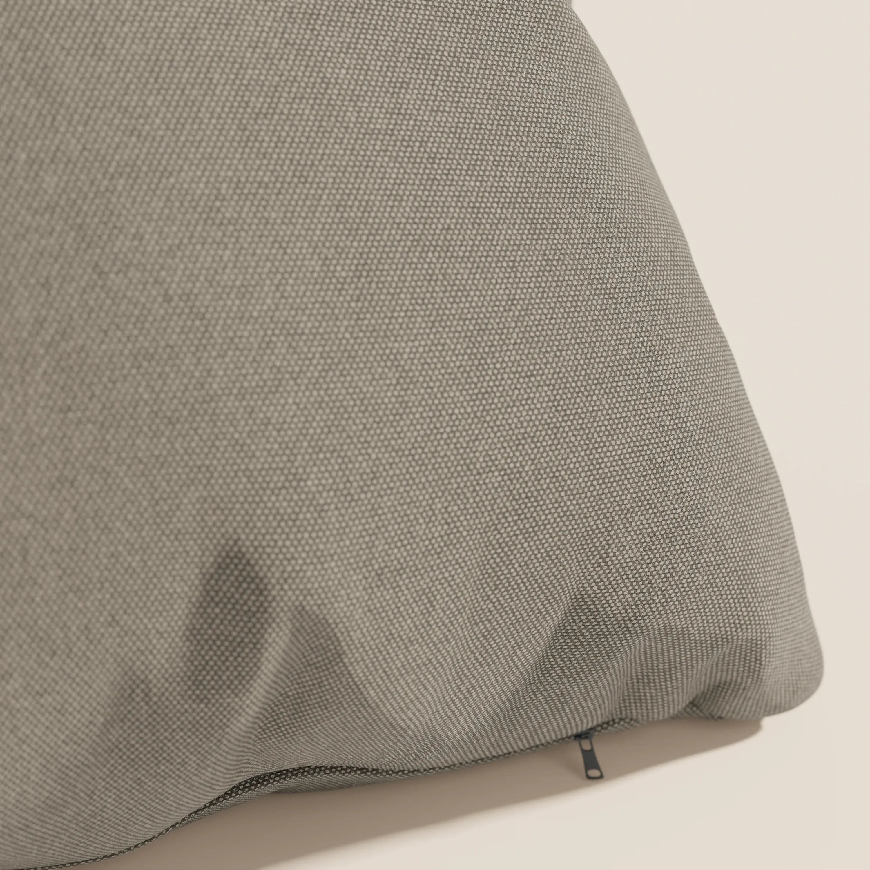 Primo piano di un cuscino Divani.Store Honey 45x45 cm in tortora T13, realizzato in tessuto impermeabile simil cotone. Le pieghe visibili, la sottile lucentezza e la cerniera nera spuntano dal bordo inferiore su uno sfondo chiaro.