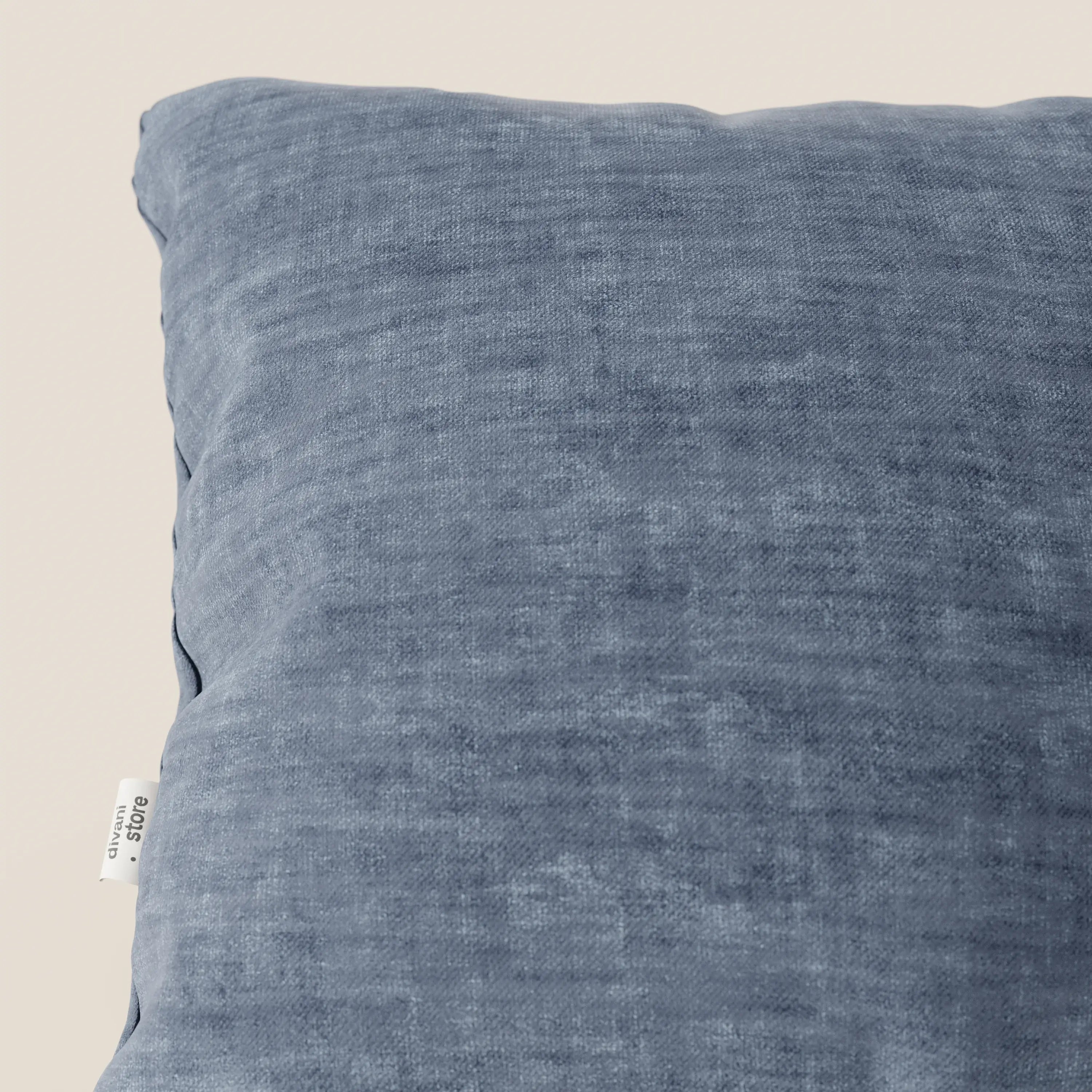 Primo piano del cuscino Sofia di Divani.Store in velluto nuvolato blu Easy Clean T35, con una superficie morbida e screziata e una piccola etichetta bianca sul bordo sinistro, su uno sfondo chiaro.