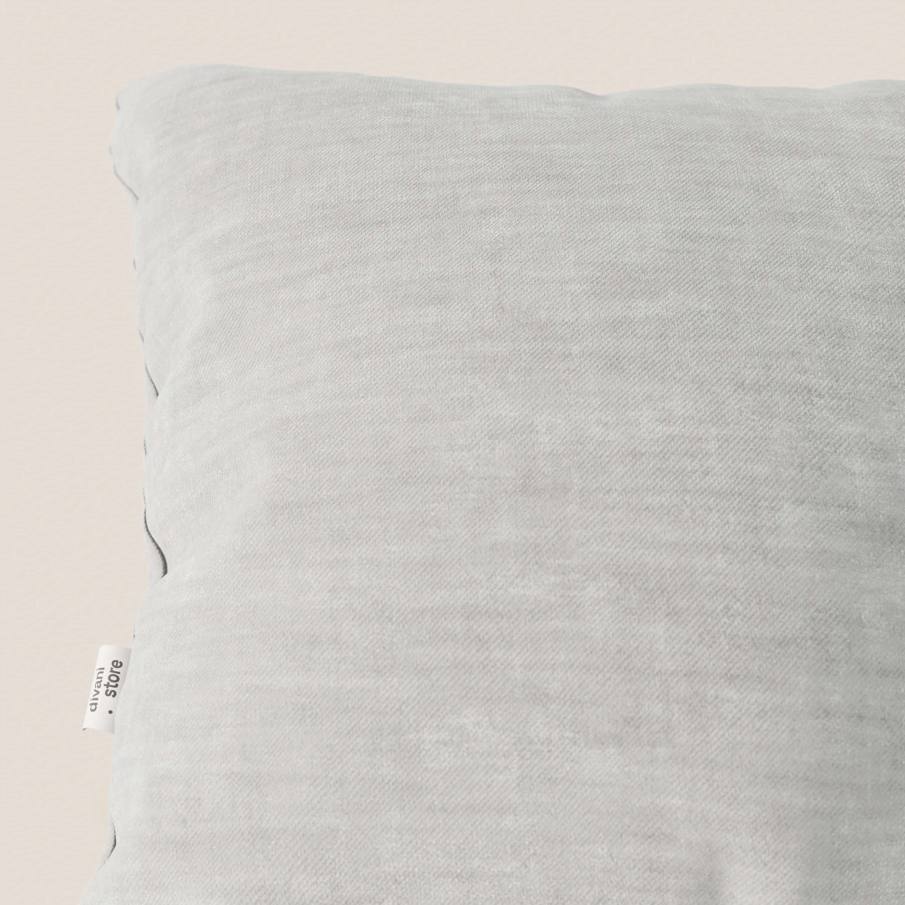 Primo piano del cuscino Sofia grigio chiaro di Divani.Store in morbido velluto Easy Clean T35, antimacchia e impermeabile. Sullo sfondo beige è visibile solo la parte superiore sinistra con l'etichetta bianca del marchio.