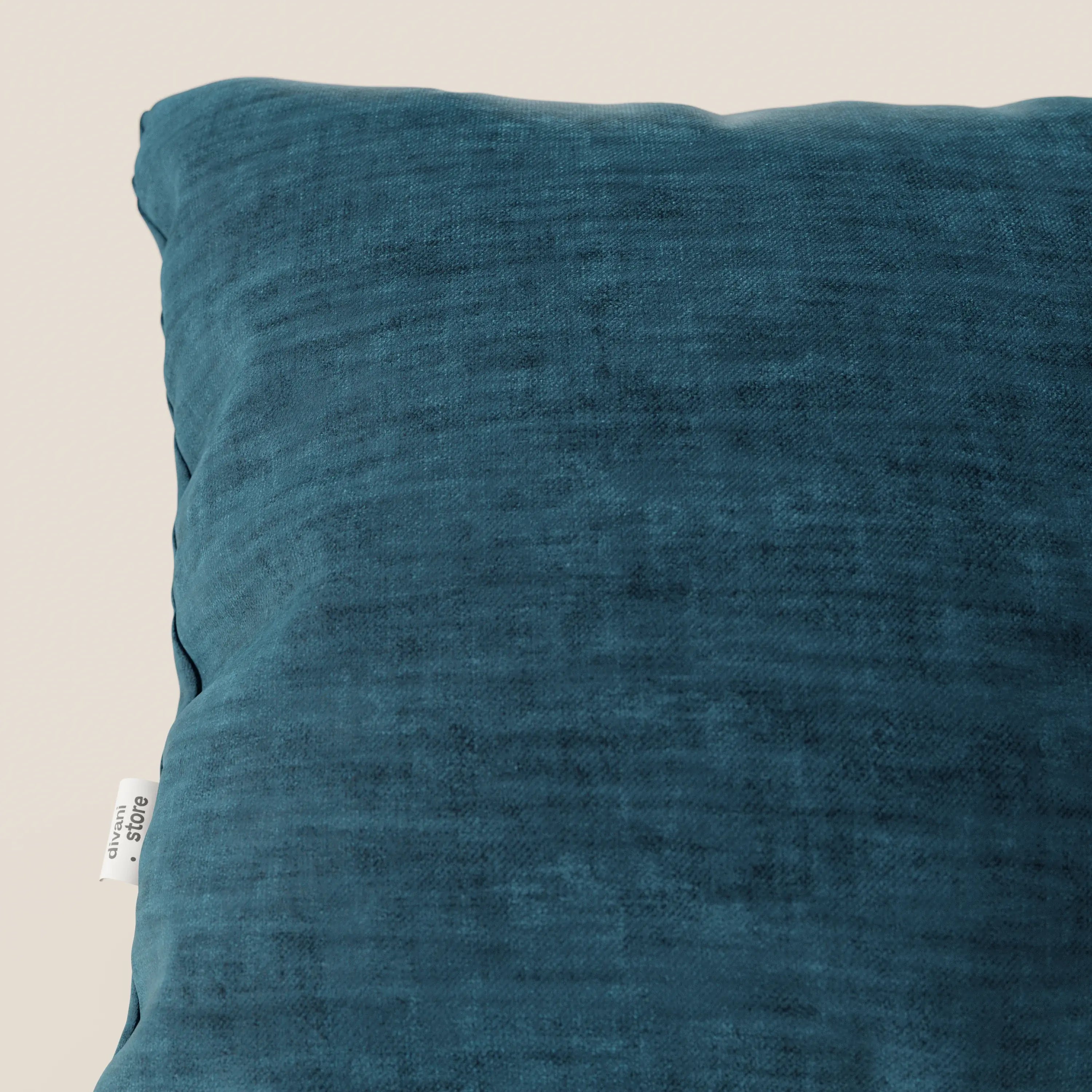 Primo piano del cuscino Sofia di Divani.Store, presentato in velluto blu acqua quadrato (morbido velluto nuvolato smacchiabile e impermeabile Easy Clean T35), posizionato in diagonale con una piccola etichetta bianca, su uno sfondo beige chiaro.