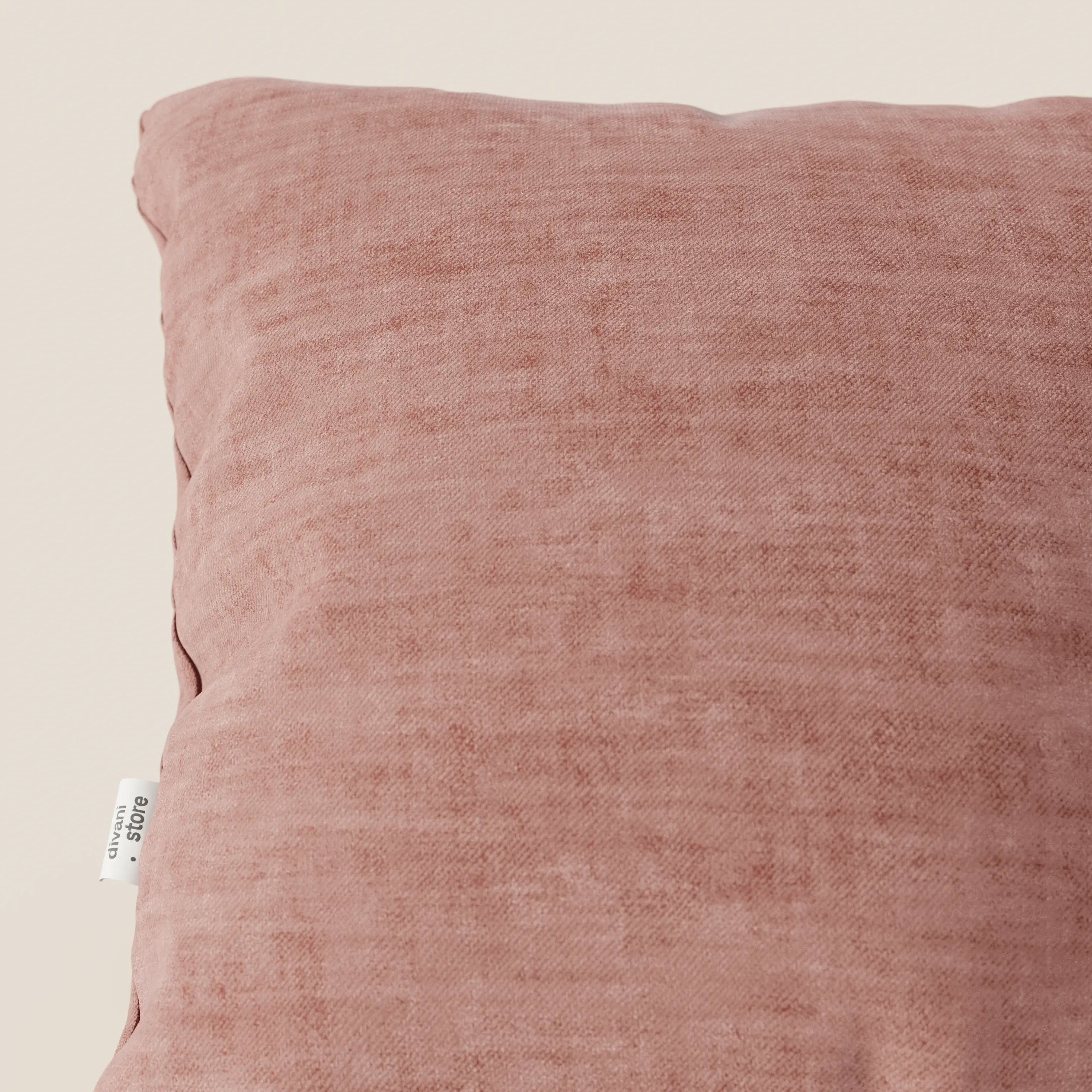 Primo piano del cuscino Sofia di Divani.Store, in velluto Easy Clean T35 rosa tenue con texture screziata e etichetta bianca sul bordo sinistro, su uno sfondo crema chiaro.