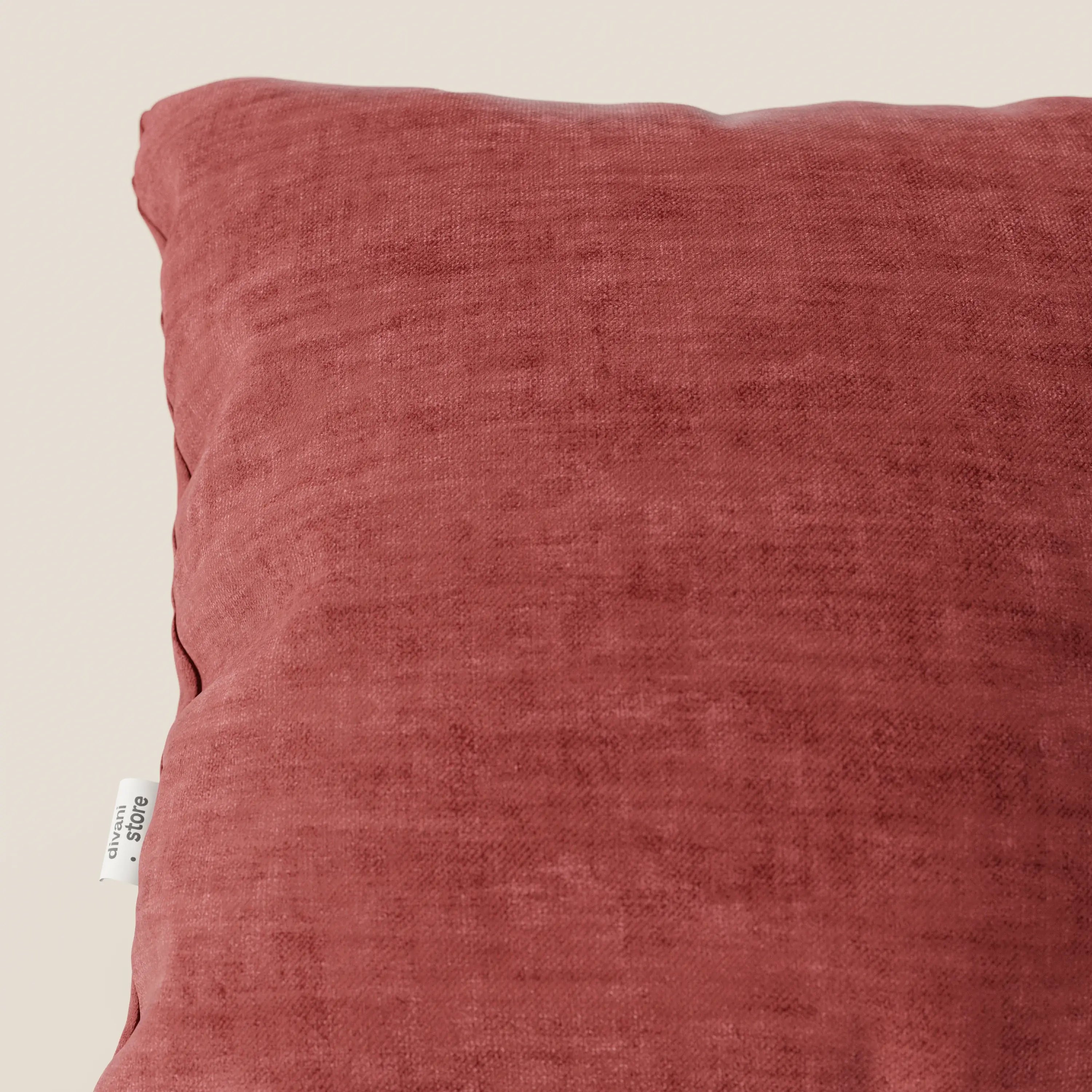 Primo piano del cuscino Sofia di Divani.Store, in velluto nuvolato Easy Clean T35 lavabile e impermeabile rosso ruggine, su uno sfondo beige tinta unita. L'etichetta del tessuto è visibile sulla cucitura sinistra; il cuscino ha una consistenza morbida e vellutata.