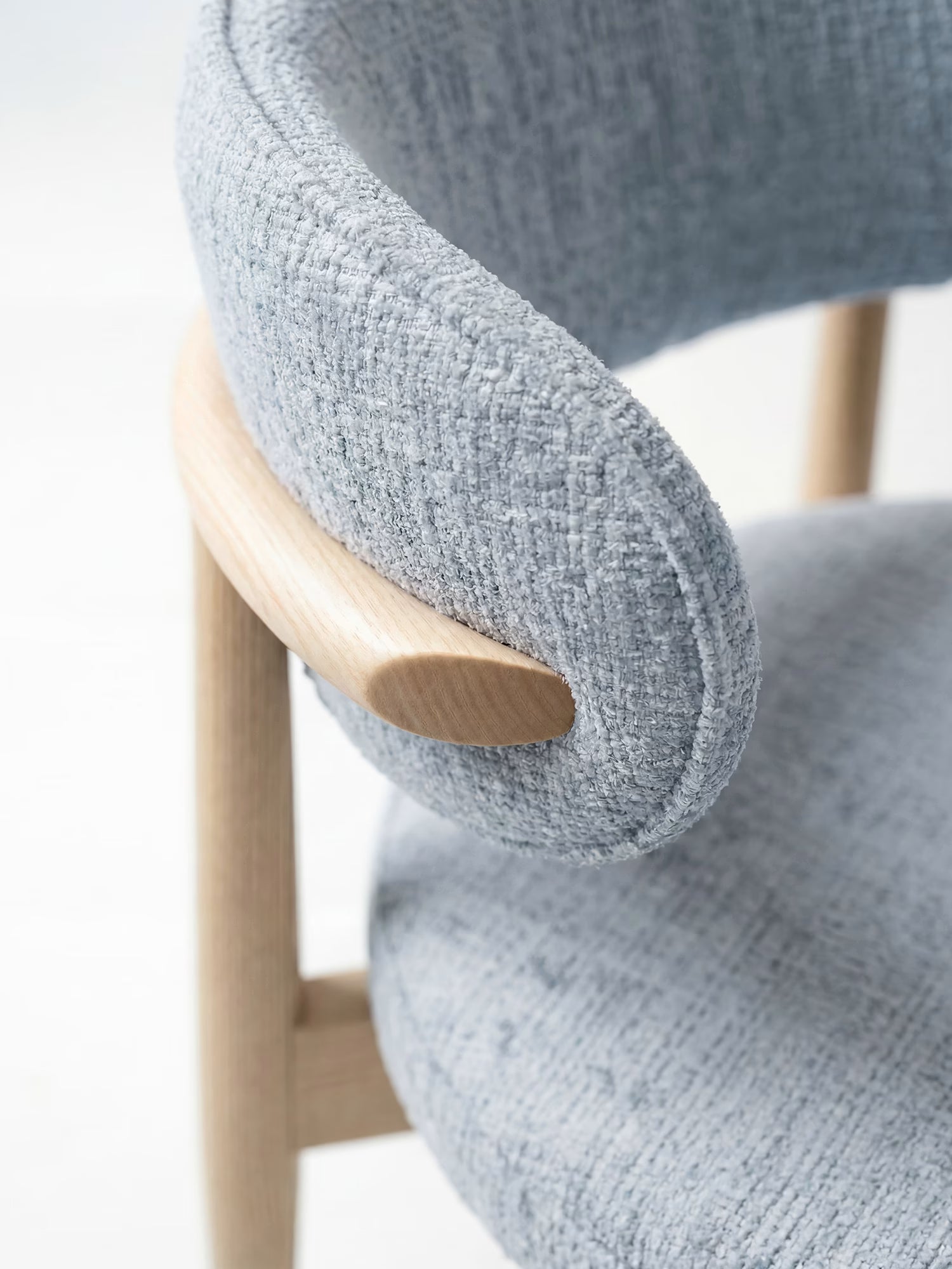 Primo piano della poltrona imbottita Dafne di Divani.Store, realizzata in tessuto ciniglia grigio chiaro certificato OEKO-TEX® e con struttura in rovere naturale massiccio, caratterizzata da uno schienale arrotondato e imbottito che le conferisce un look minimalista di ispirazione scandinava.