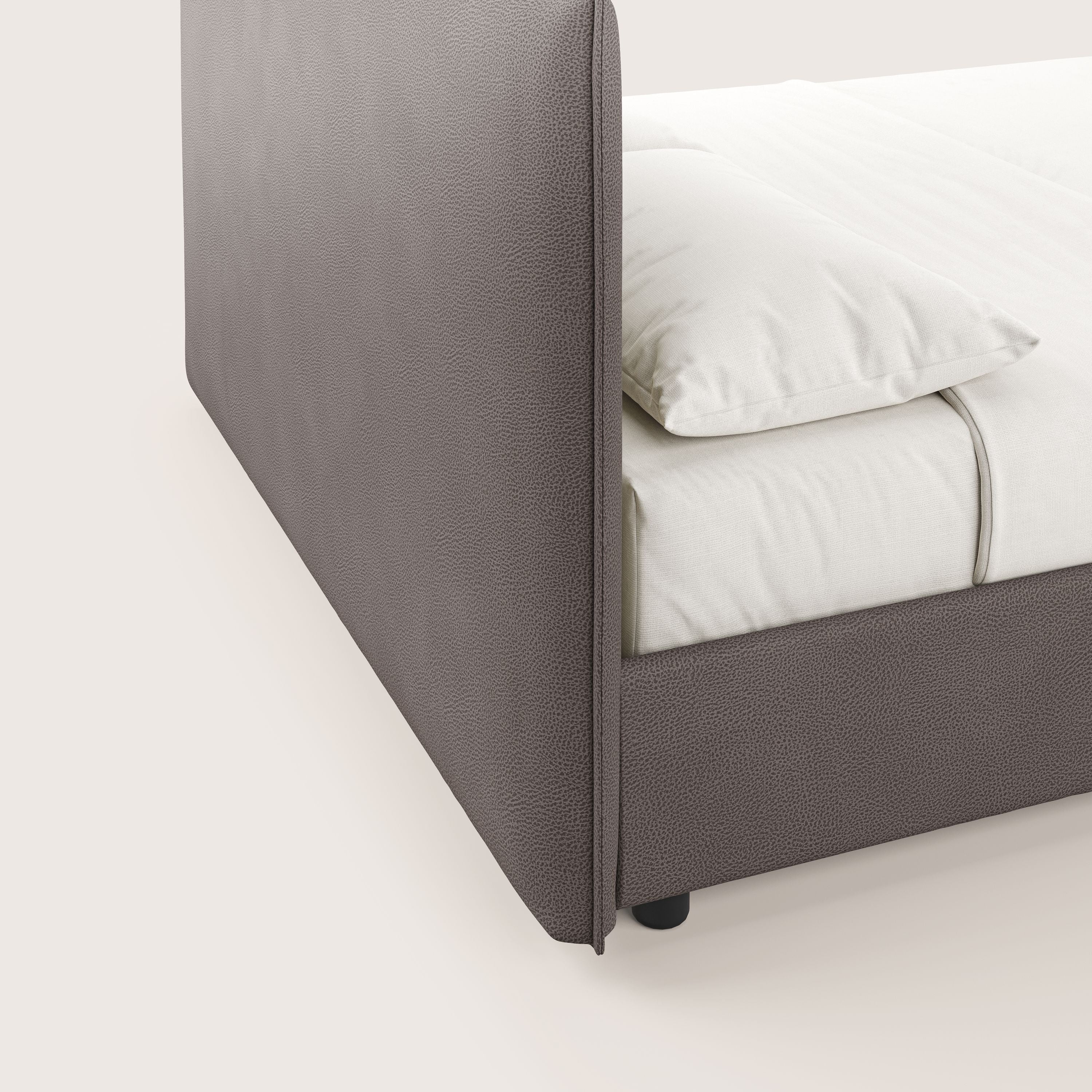 Primo piano del letto contenitore Nina di Letti.store, caratterizzato da un'alta testiera imbottita in microfibra marrone antimacchia e da un giroletto minimal coordinato. Realizzato in modo ordinato con biancheria bianca, le gambe nere sono visibili su uno sfondo semplice.