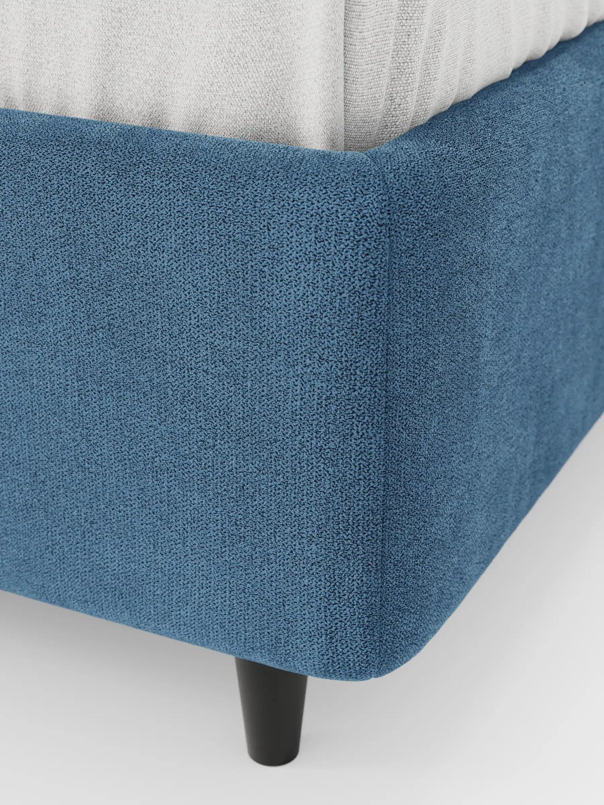 Un primo piano dell'angolo di un letto sommier imbottito ERON con vano contenitore in tessuto impermeabile certificato Oeko-Tex, caratterizzato da un tessuto blu scuro a trama rigata, una gamba cilindrica corta di colore scuro e un materasso di colore chiaro appoggiato sopra.