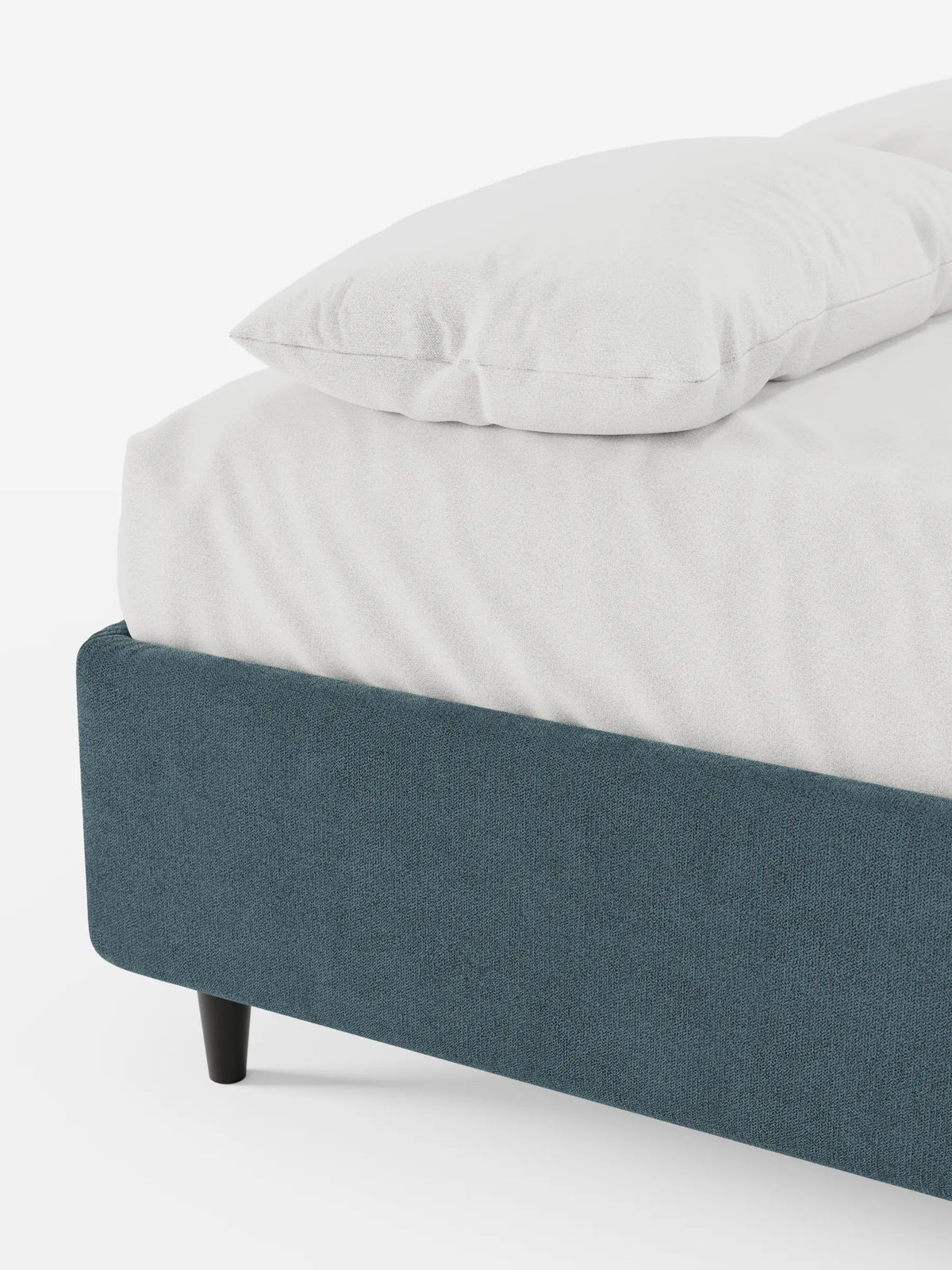 Vista angolare di un letto sommier imbottito ERON con vano contenitore in tessuto impermeabile certificato Oeko-Tex di colore blu intenso, rivestito con biancheria da letto grigio chiaro, su gambe scure e affusolate, su uno sfondo bianco.