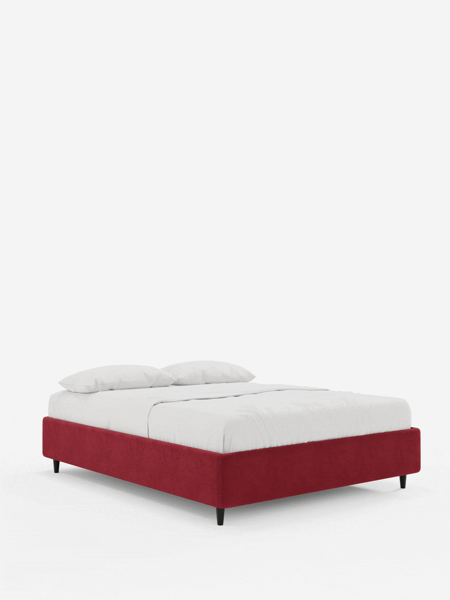 Un letto sommier imbottito ERON con vano contenitore in tessuto impermeabile certificato Oeko-Tex color rosso intenso, ripreso da un’angolazione bassa con base rettangolare, gambe corte nere, un materasso bianco candido e due cuscini su uno sfondo a tinta unita.