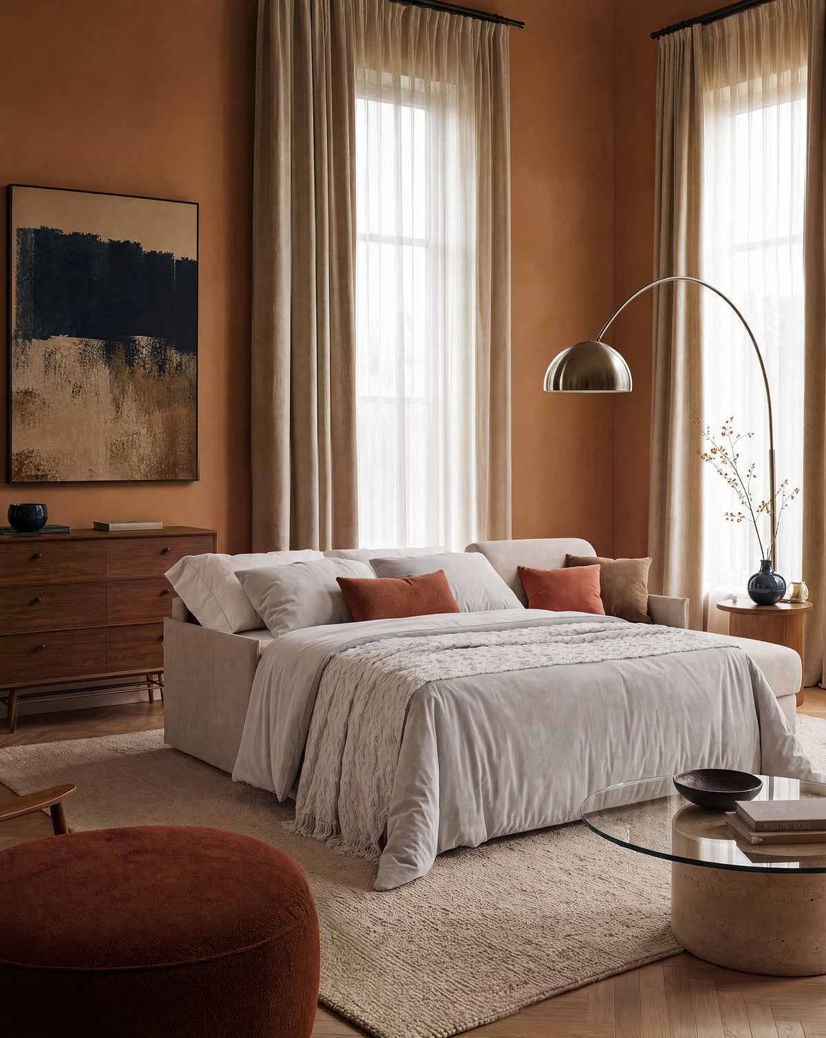 Un'accogliente camera da letto con pareti color cuoio e ampie finestre adornate da tende trasparenti beige. Lo spazio è arredato con il divano letto angolare con penisola contenitore Evans di Divani.Store, un divano letto di design dalle dimensioni compatte rivestito in tessuto T23 facile da pulire, impreziosito da un piumone grigio chiaro e da cuscini color ruggine. A completare l’arredamento della stanza ci sono un comò in legno, un quadro astratto, una moderna lampada da terra ad arco e un tavolino