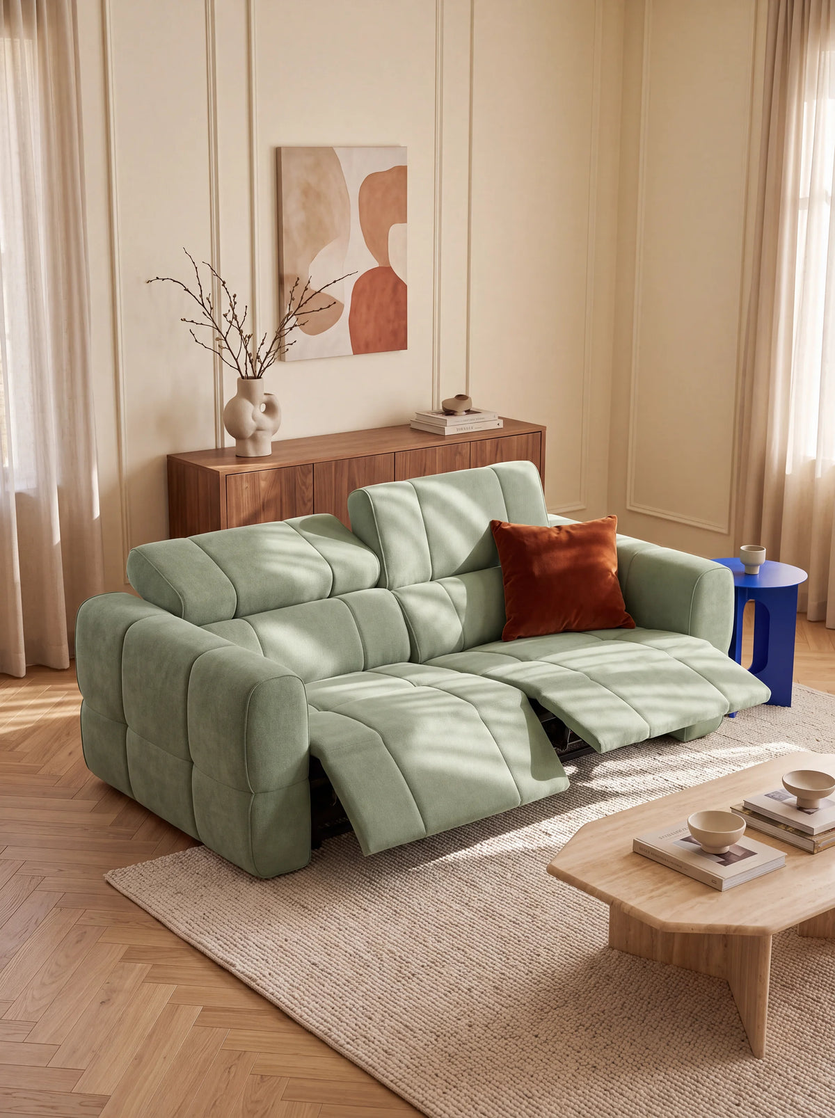 Un divano modulare Giotto a 3 posti color verde salvia, con poggiapiedi reclinabile, cuscini con cuciture a griglia, poggiatesta reclinabili e un cuscino decorativo arancione, è posizionato su un tappeto chiaro tra un tavolino da caffè in legno e un tavolino blu.
