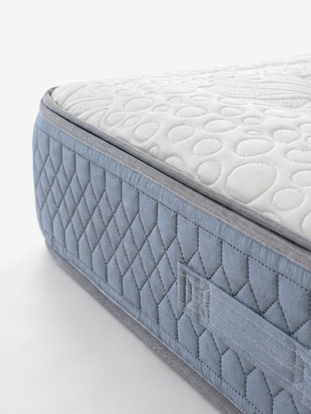 Primo piano dell'angolo del KYROS – Materasso Luxury di Divani.Store, caratterizzato da una parte superiore bianca con motivi circolari e da un lato blu trapuntato, impreziosito da cuciture a rombi e da una maniglia in tessuto. L’immagine mette in risalto la trama dettagliata e i bordi ben definiti su uno sfondo bianco, evidenziando l’avanzata struttura di sostegno con micro molle insacchettate del materasso e il design a 7 zone con supporto posturale intelligente.