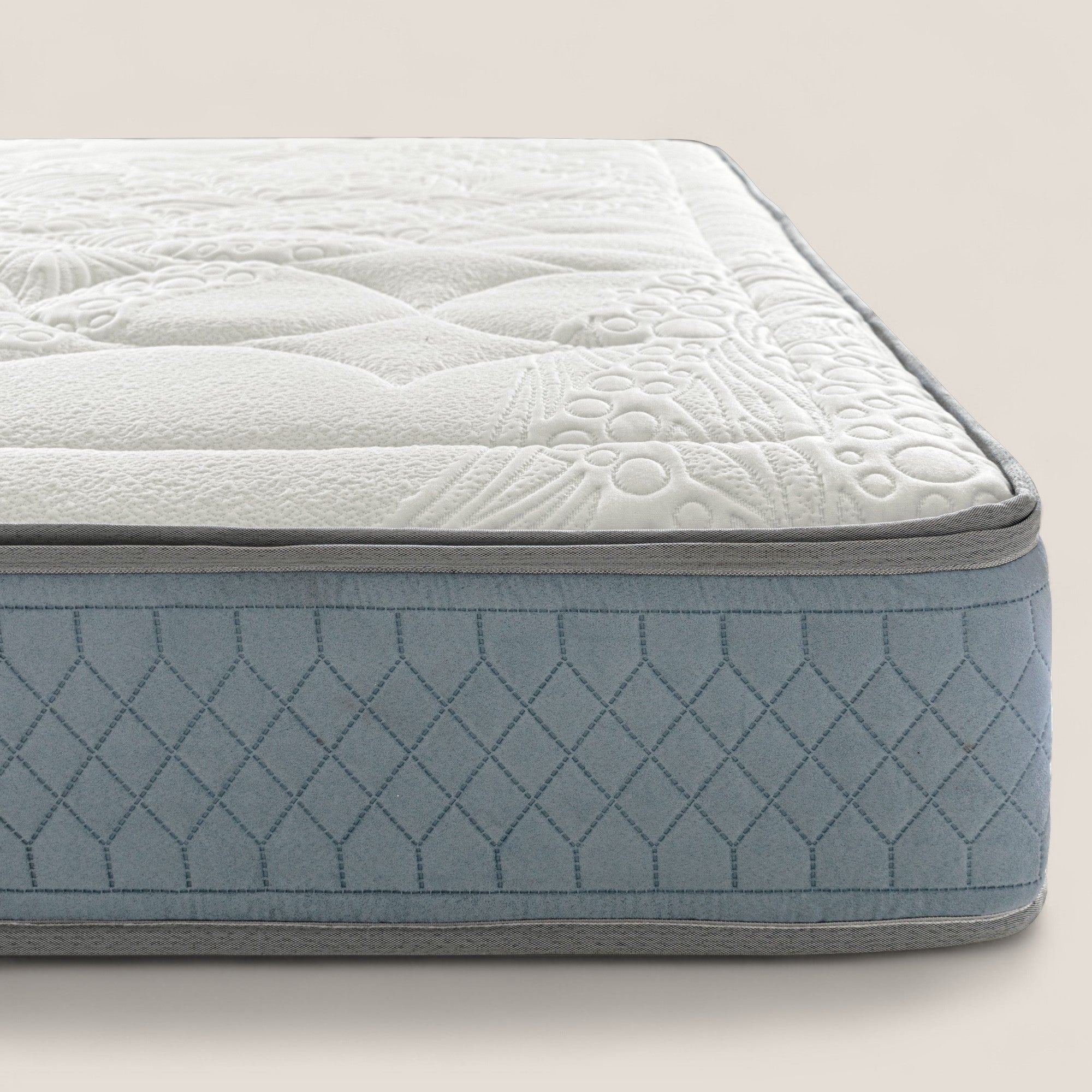 Un primo piano del materasso KYROS di Divani.Store mostra il top bianco con cuciture floreali e circolari, i fianchi trapuntati a rombi azzurri, i bordi grigi e il comfort stratificato della schiuma di memoria e delle molle insacchettate a 7 zone.