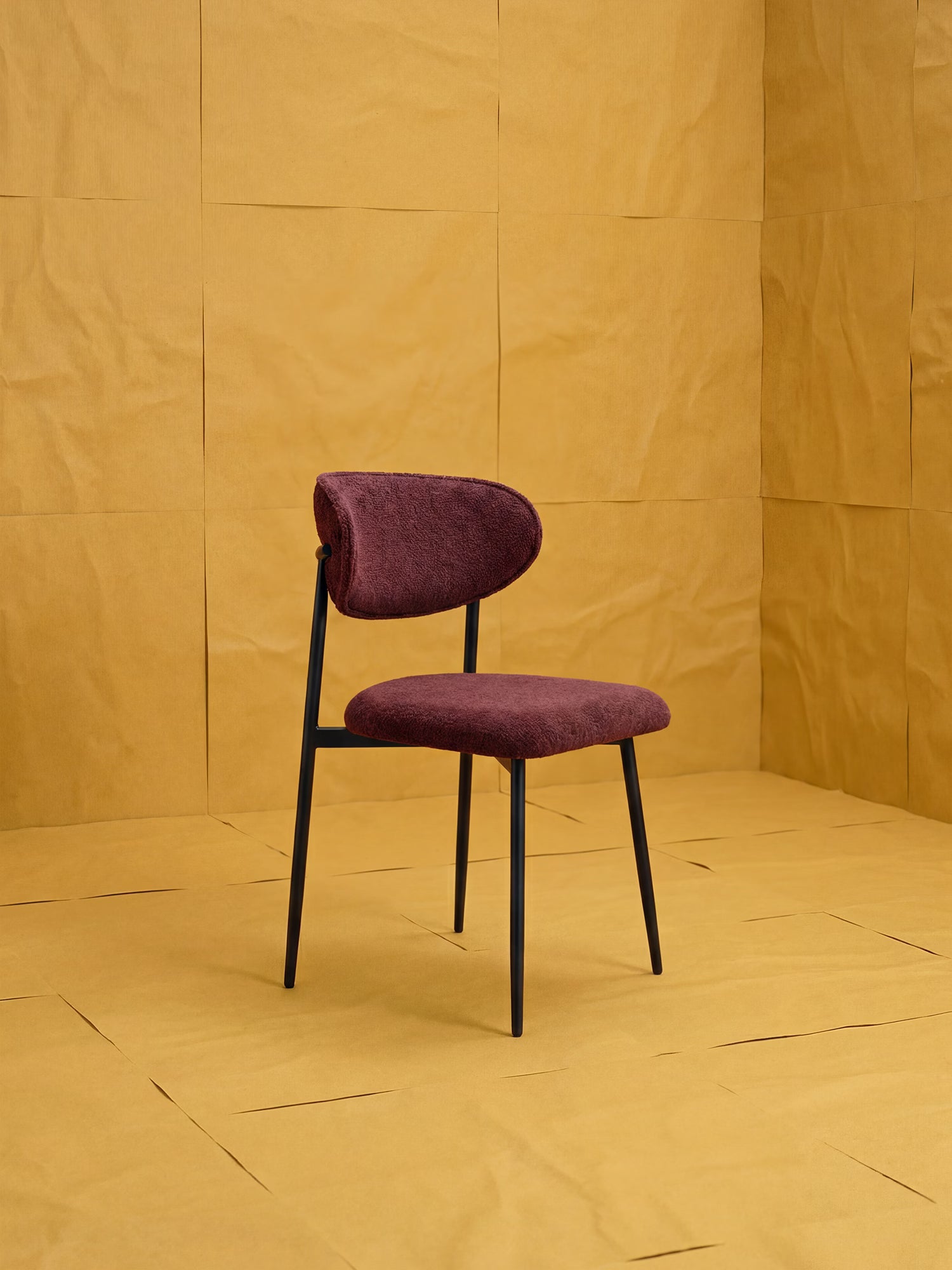 La Luce, una sedia imbottita in morbido tessuto ciniglia bordeaux di Divani.Store, con struttura in metallo nero, si trova in una stanza decorata con fogli di carta giallo senape disposti a griglia sulle pareti e sul pavimento.