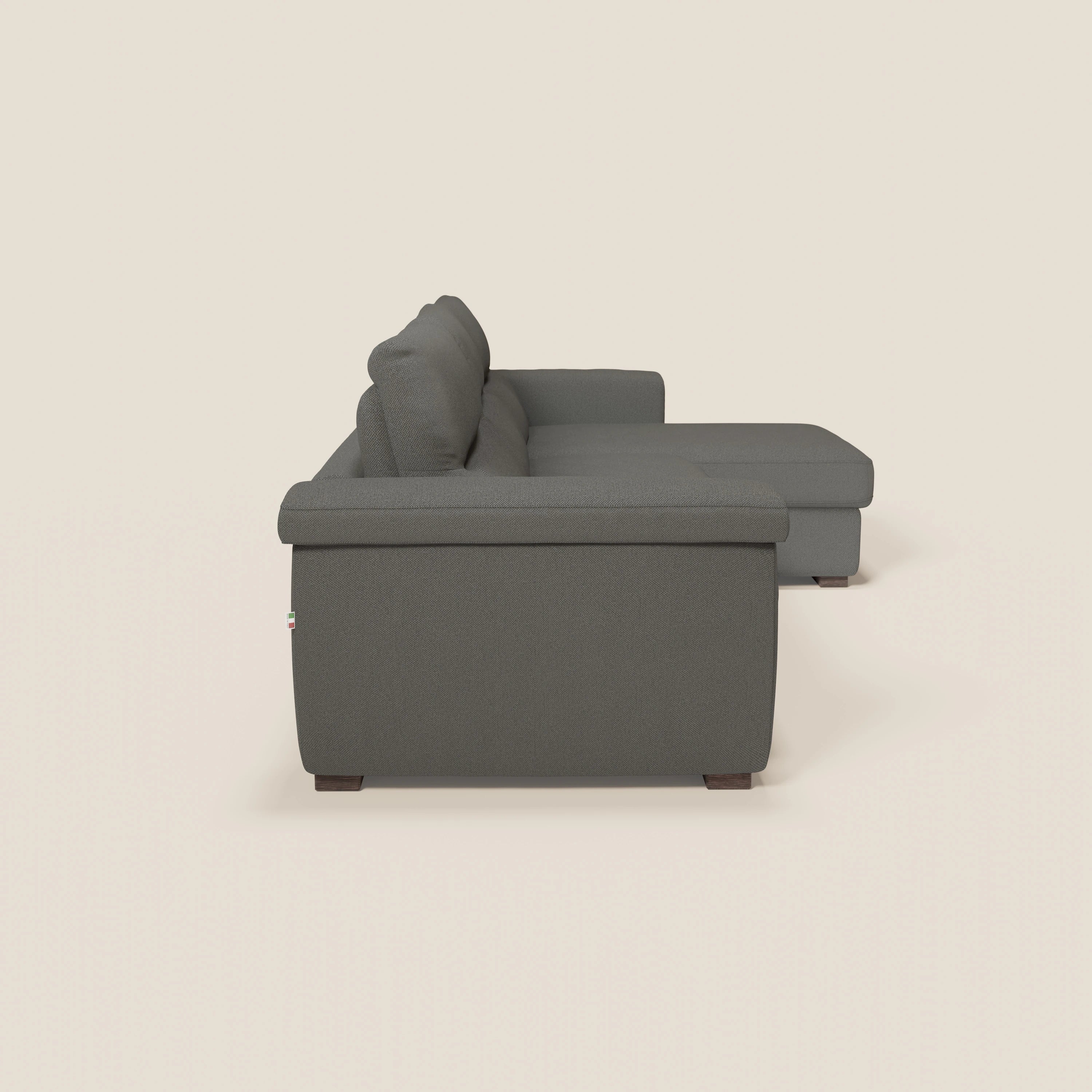 Un divano letto angolare Magnolia con penisola contenitore in tessuto smacchiabile T05 antracite di Divani.Store è rappresentato lateralmente su sfondo beige chiaro, con un ampio bracciolo, materasso H18 cm e cuscini spessi.