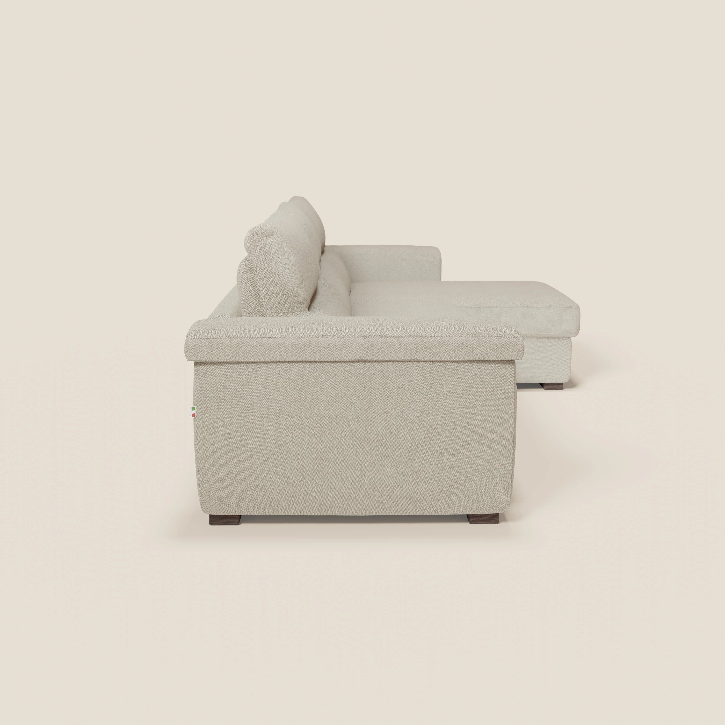 Vista laterale del divano letto angolare Magnolia con Penisola Contenitore di Divani.Store in tessuto smacchiabile T05 panna, materasso H18 cm, cuscini in peluche, gambe basse in legno scuro, presentato su uno sfondo bianco sporco.