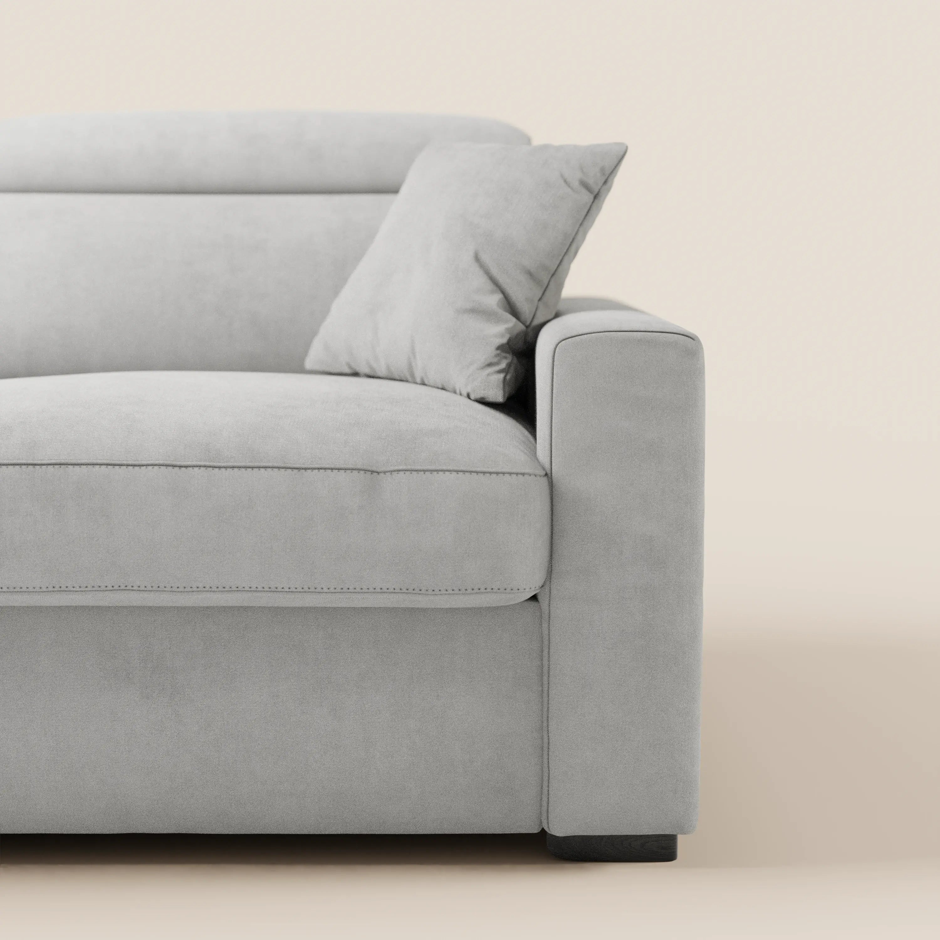 Il divano letto a 3 posti Maximus con materasso ALTO 20 cm di Divani.Store è presentato in grigio chiaro con linee pulite, braccioli rettangolari, cuscini morbidi, un cuscino coordinato e poggiatesta reclinabili, su uno sfondo beige a tinta unita.