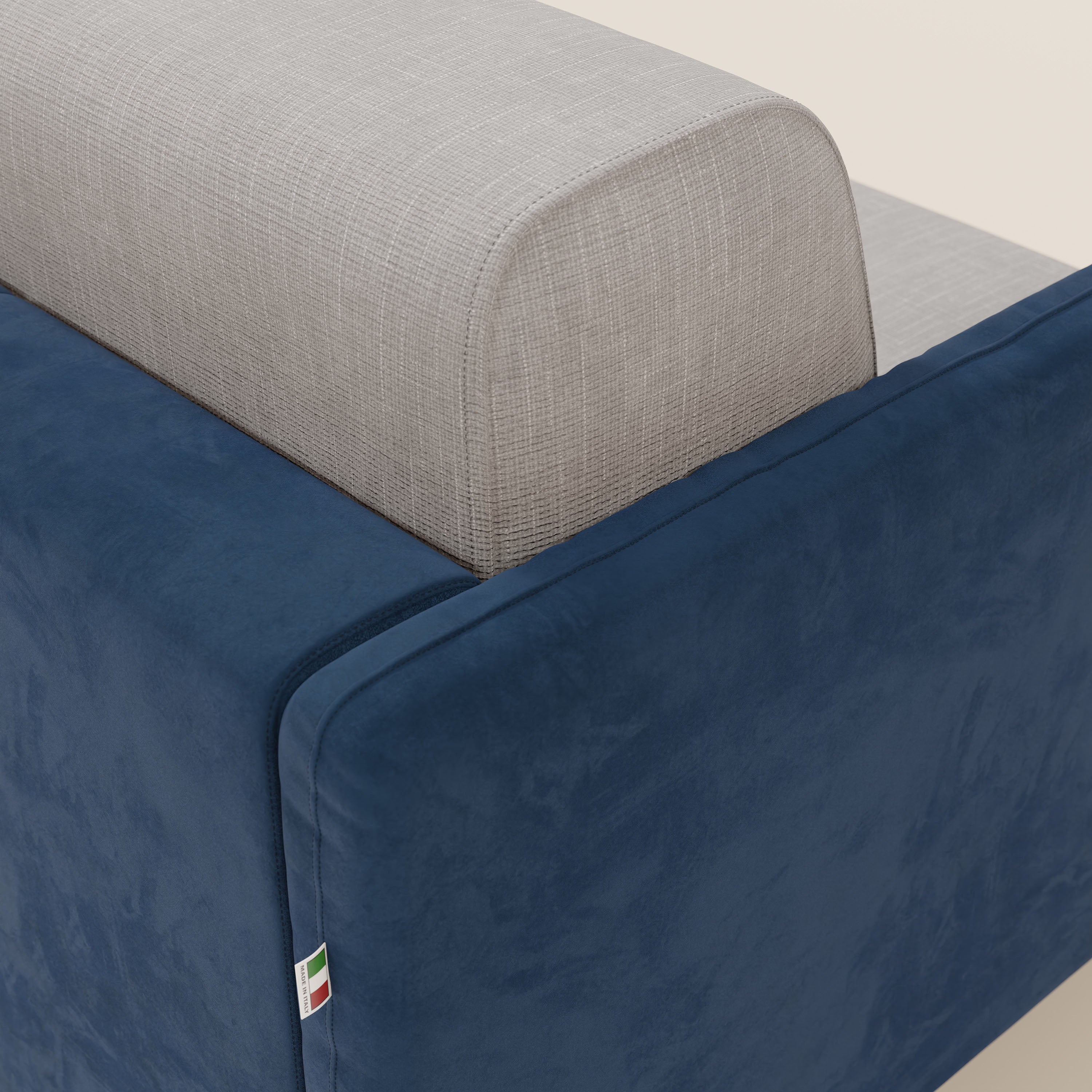 Primo piano del divano letto Narciso di Divani.Store: schienale grigio chiaro strutturato ed esterno blu intenso liscio in microfibra antimacchia e tessuto impermeabile T06. Una targhetta con i colori della bandiera italiana è cucita vicino al fondo su uno sfondo neutro.