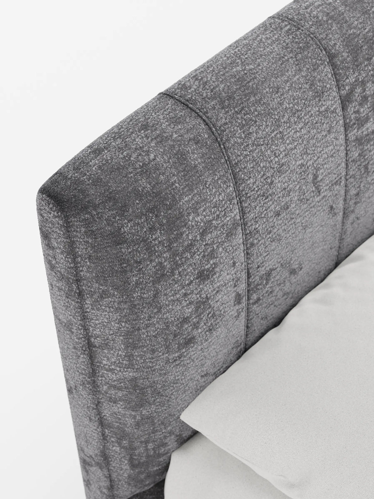 Un primo piano della testiera grigio scuro, con trapuntatura a canali verticali, del letto imbottito ONYX con contenitore e testiera verticale in tessuto certificato Oeko-Tex®, con un cuscino grigio chiaro appoggiato contro di essa su uno sfondo bianco.