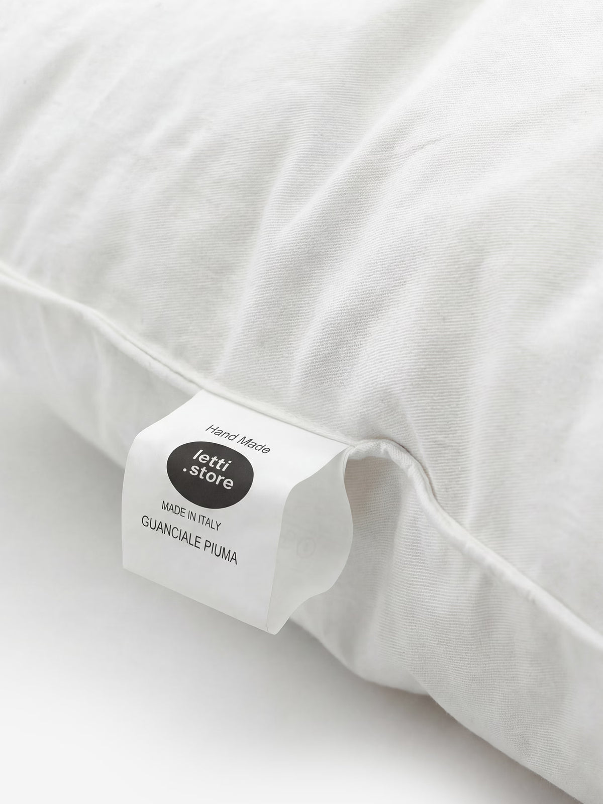 Un primo piano di un cuscino bianco con un’etichetta in tessuto su cui si leggono le scritte “Divani.Store”, “Hand Made”, “MADE IN ITALY” e “VELTHA-ORGANIC Guanciale in Piuma d’Oca Naturale e Certificata” su uno sfondo bianco a tinta unita. Il tessuto appare morbido e soffice, a sottolineare la qualità eccellente di questo guanciale in piuma d’oca naturale certificata, realizzato con maestria per garantire il massimo comfort.