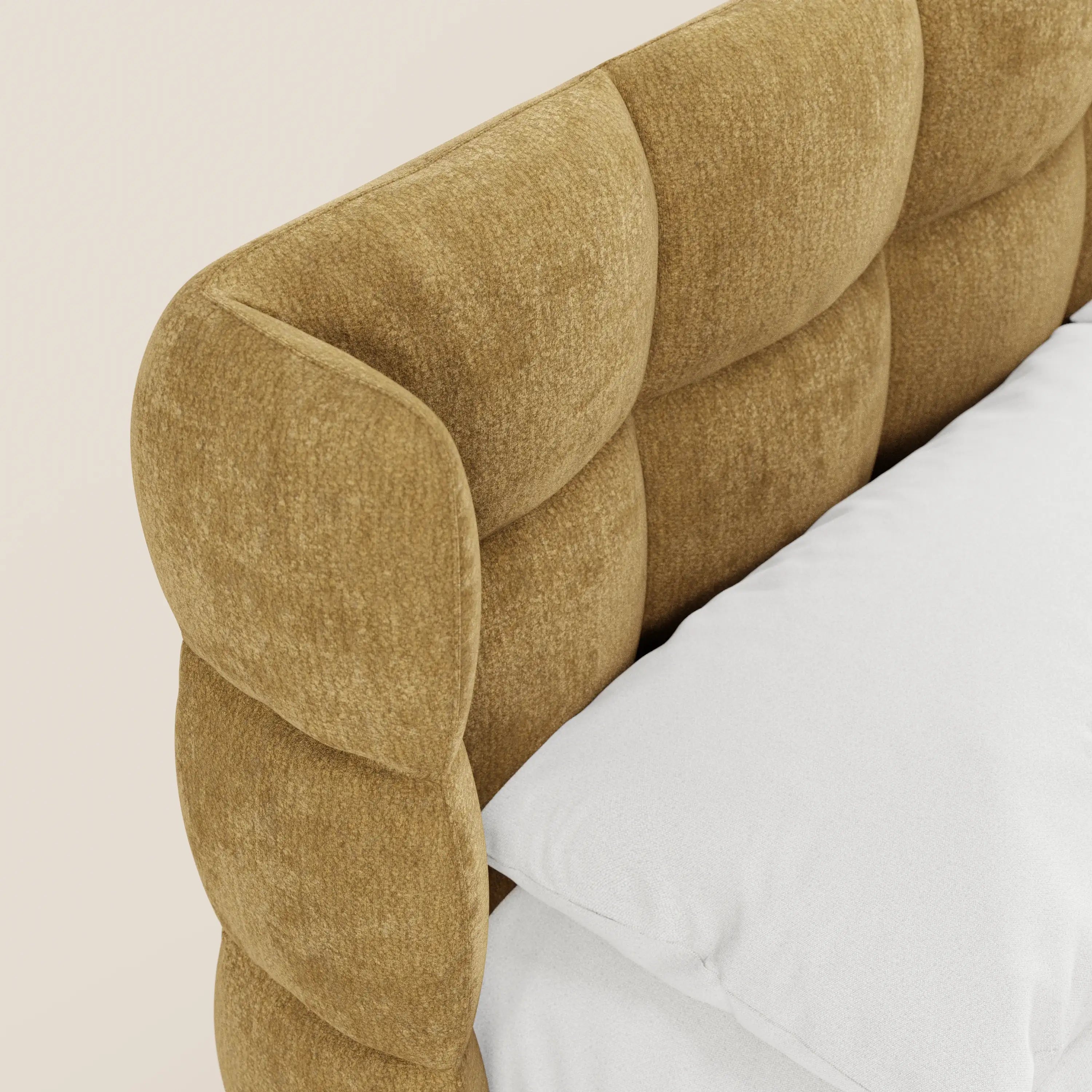 Primo piano del letto imbottito RAGNAR con contenitore e testiera capitonné avvolgente di Letti.store, realizzato in un elegante tessuto certificato Oeko-Tex®. Il materasso e i cuscini bianchi creano un contrasto con la calda struttura color marrone dorato su uno sfondo beige chiaro.