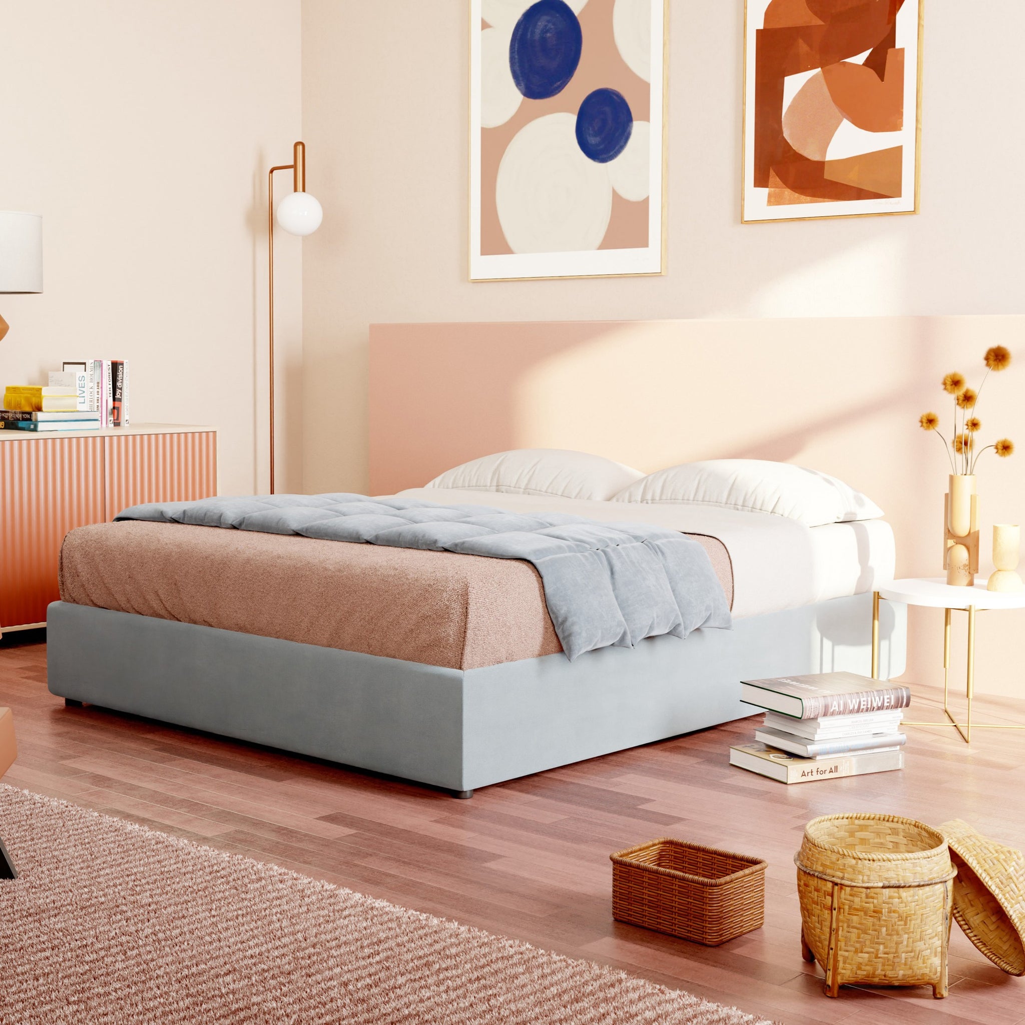 Una camera da letto accogliente con un letto imbottito azzurro, l'Ina sommier con ampio contenitore di Letti.store, cuscini bianchi e una coperta beige trasuda design minimalista. L'arte astratta adorna le pareti accanto a un'alta lampada da terra bianca e una cassettiera in legno. Pavimenti illuminati dal sole e cestini di vimini aggiungono serenità.