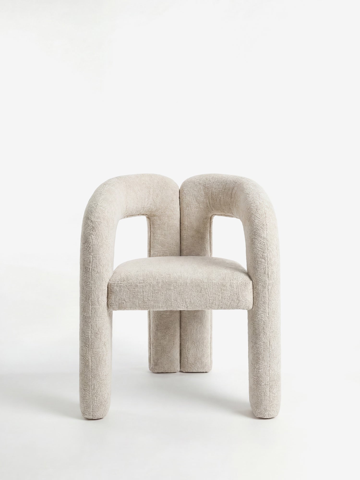 La sedia di design Sibilla di Divani.Store presenta gambe spesse e arrotondate e uno schienale ricurvo, il tutto interamente rivestito in tessuto beige chiaro. Le sue forme tubolari decise e la linea arcuata creano un effetto scultoreo sullo sfondo bianco a tinta unita.