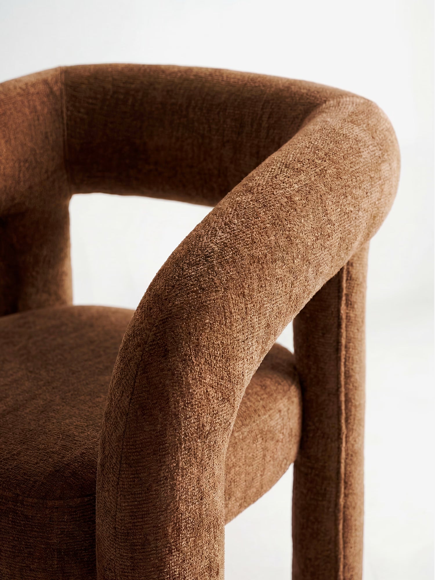 Primo piano della sedia di design “Sibilla” di Divani.Store, rivestita in tessuto marrone testurizzato con struttura interamente rivestita e forma arcuata, caratterizzata da schienale arrotondato e braccioli che si incurvano verso le gambe cilindriche su uno sfondo chiaro.