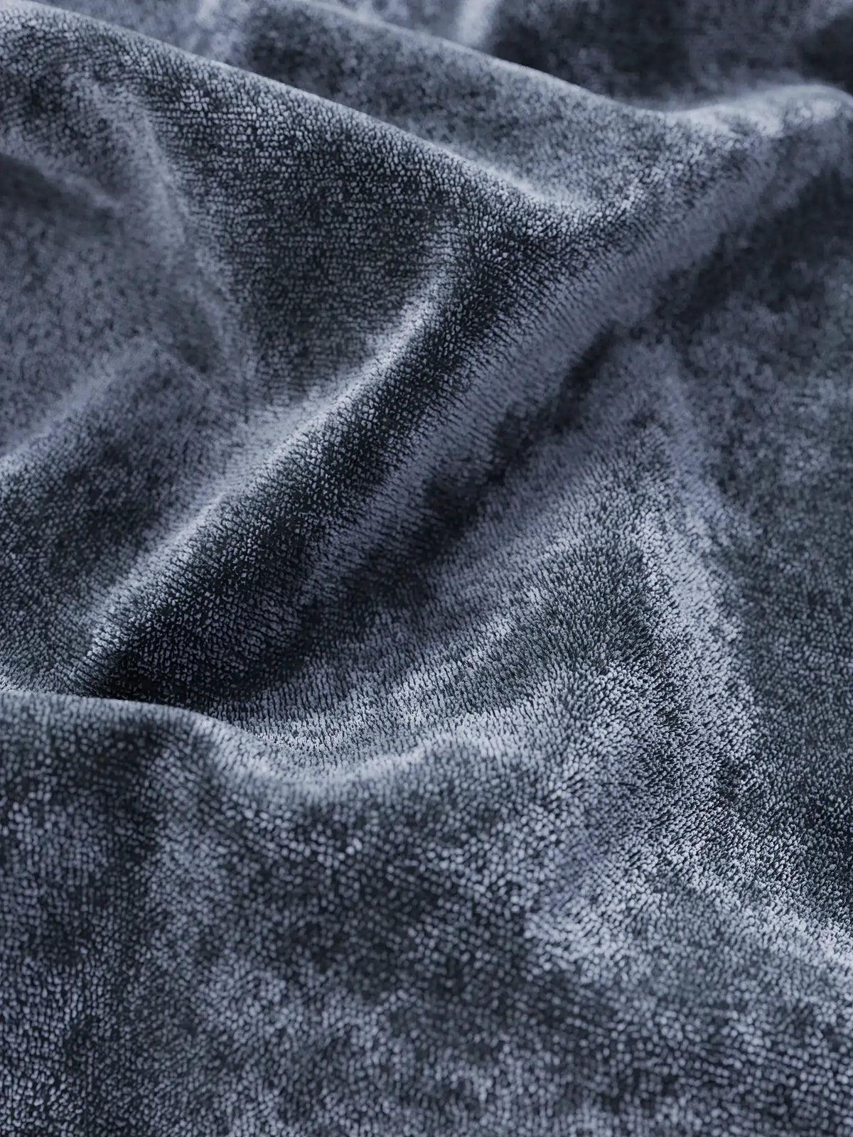 Primo piano del letto imbottito AURO in velluto ciniglia grigio scuro, morbido e lussuoso, con vano contenitore e testiera capitonné alta in ciniglia Oeko-Tex®, caratterizzato da pieghe profonde e morbide e da una trama che riflette la luce, esaltandone la profondità vellutata.