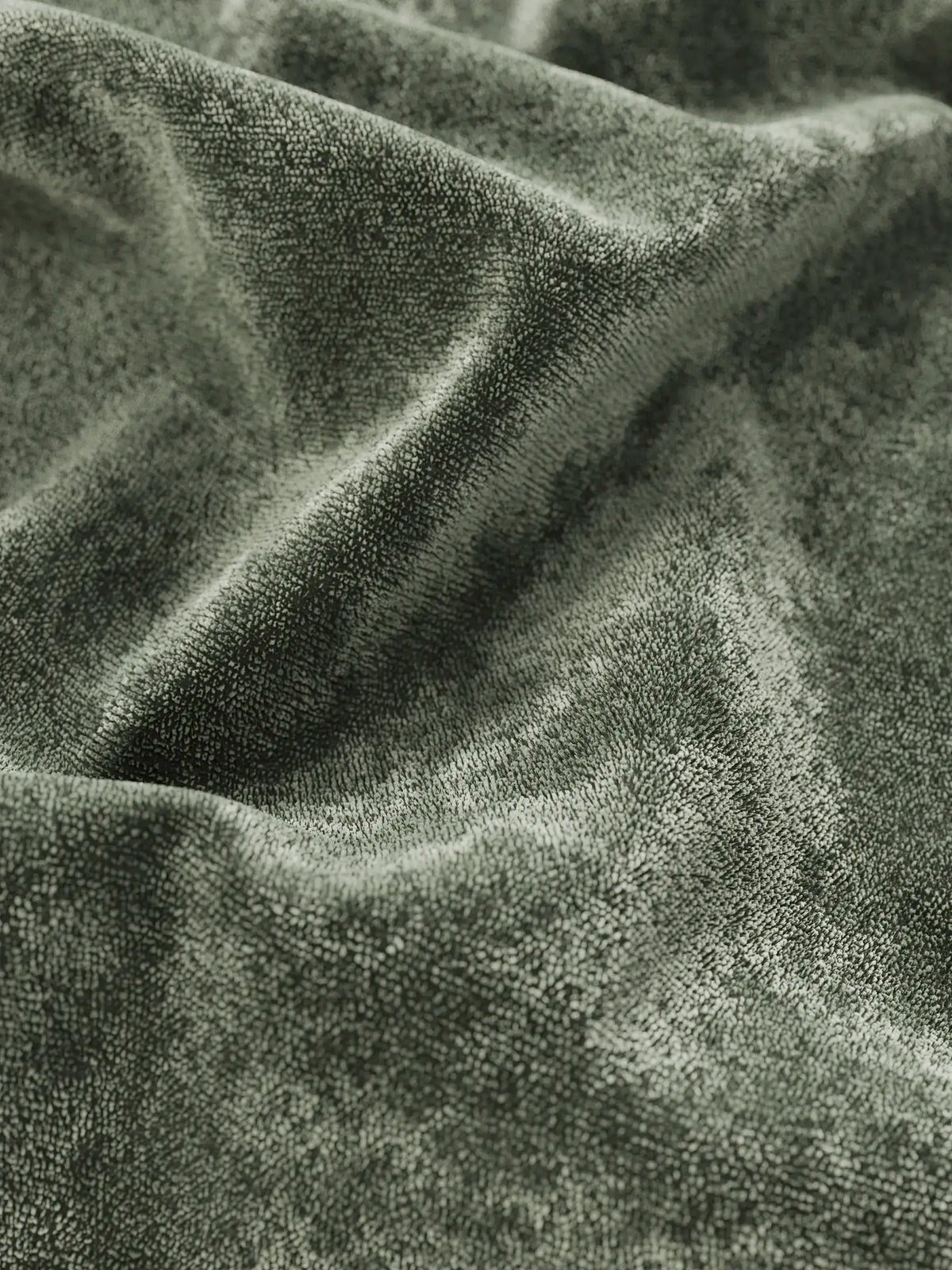 Un primo piano di un tessuto in velluto verde salvia, drappeggiato in pieghe profonde e giochi d’ombra, su un letto imbottito AURO con contenitore e testiera alta capitonné in ciniglia Oeko-Tex®, che crea una superficie morbida e ricca di texture.