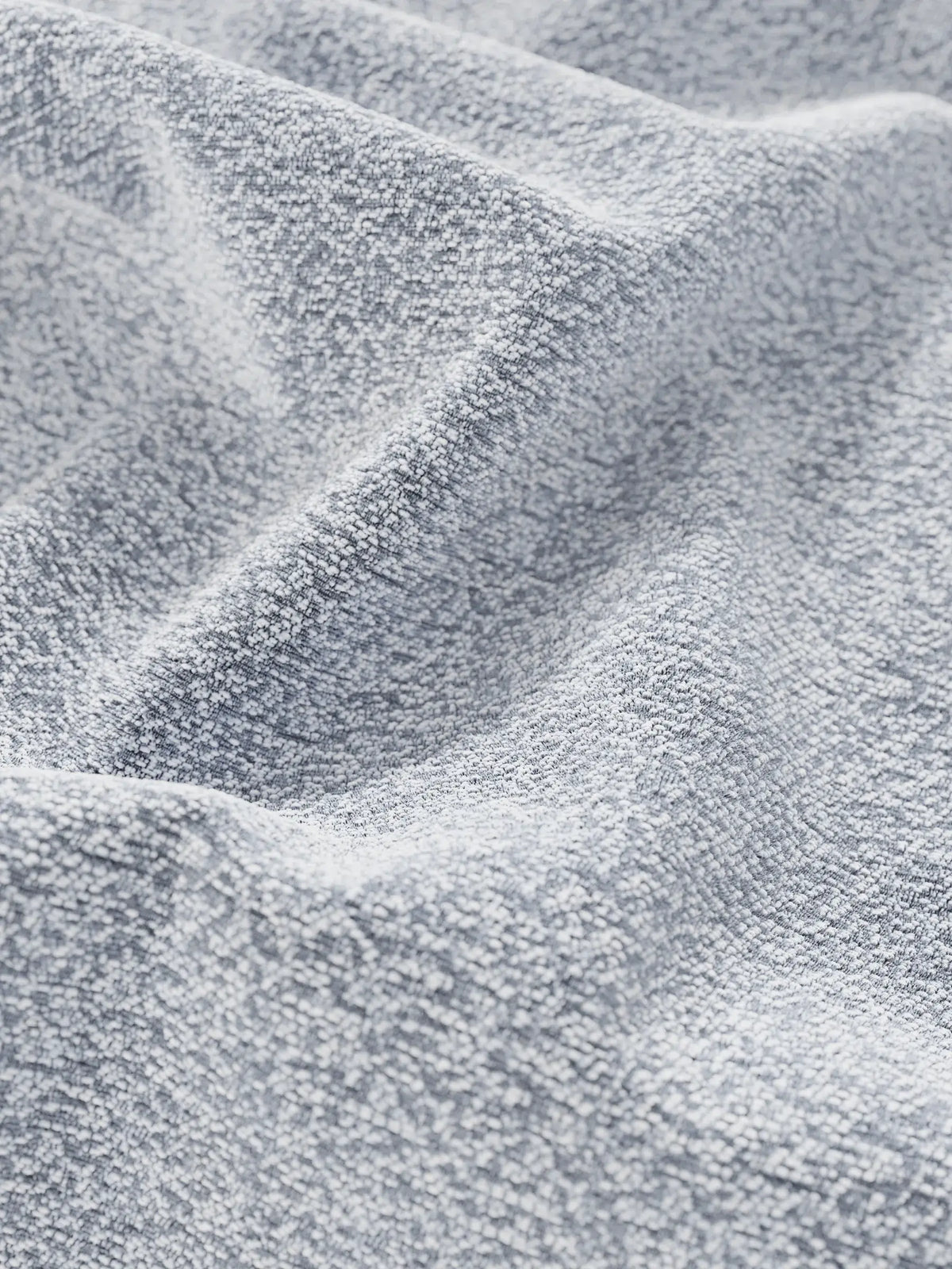 Un primo piano del tessuto intrecciato grigio chiaro e dalla trama ruvida del letto di design VALKIRIA con vano contenitore e testiera avvolgente, raccolto in morbide pieghe che mettono in risalto la profondità del materiale e la sua trama granulosa.