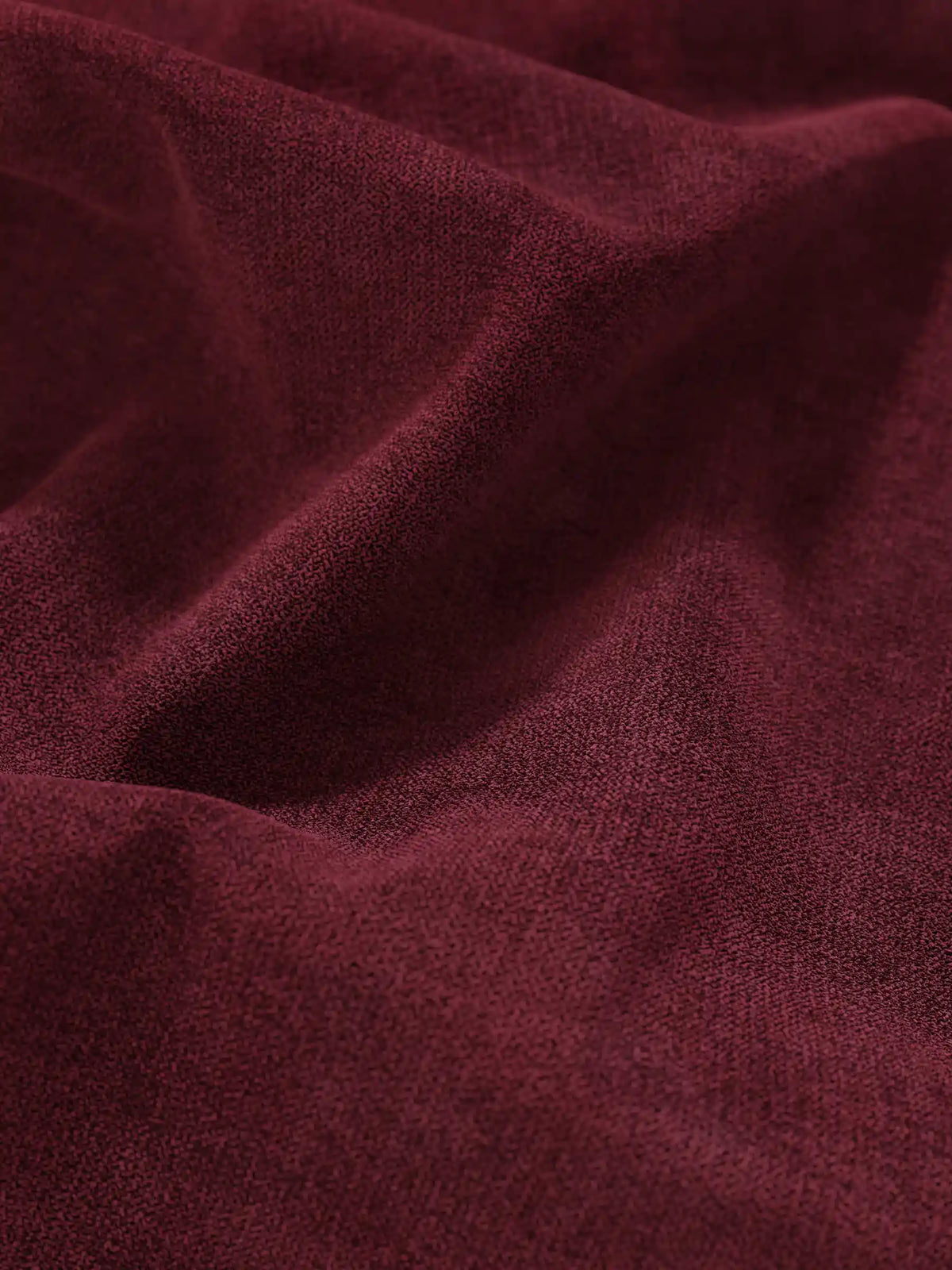 Primo piano sul tessuto color bordeaux scuro e dalla trama fine di un letto sommier imbottito ERON con vano contenitore in tessuto impermeabile certificato Oeko-Tex, drappeggiato in morbide pieghe che creano sottili giochi di luce e ombre profonde.
