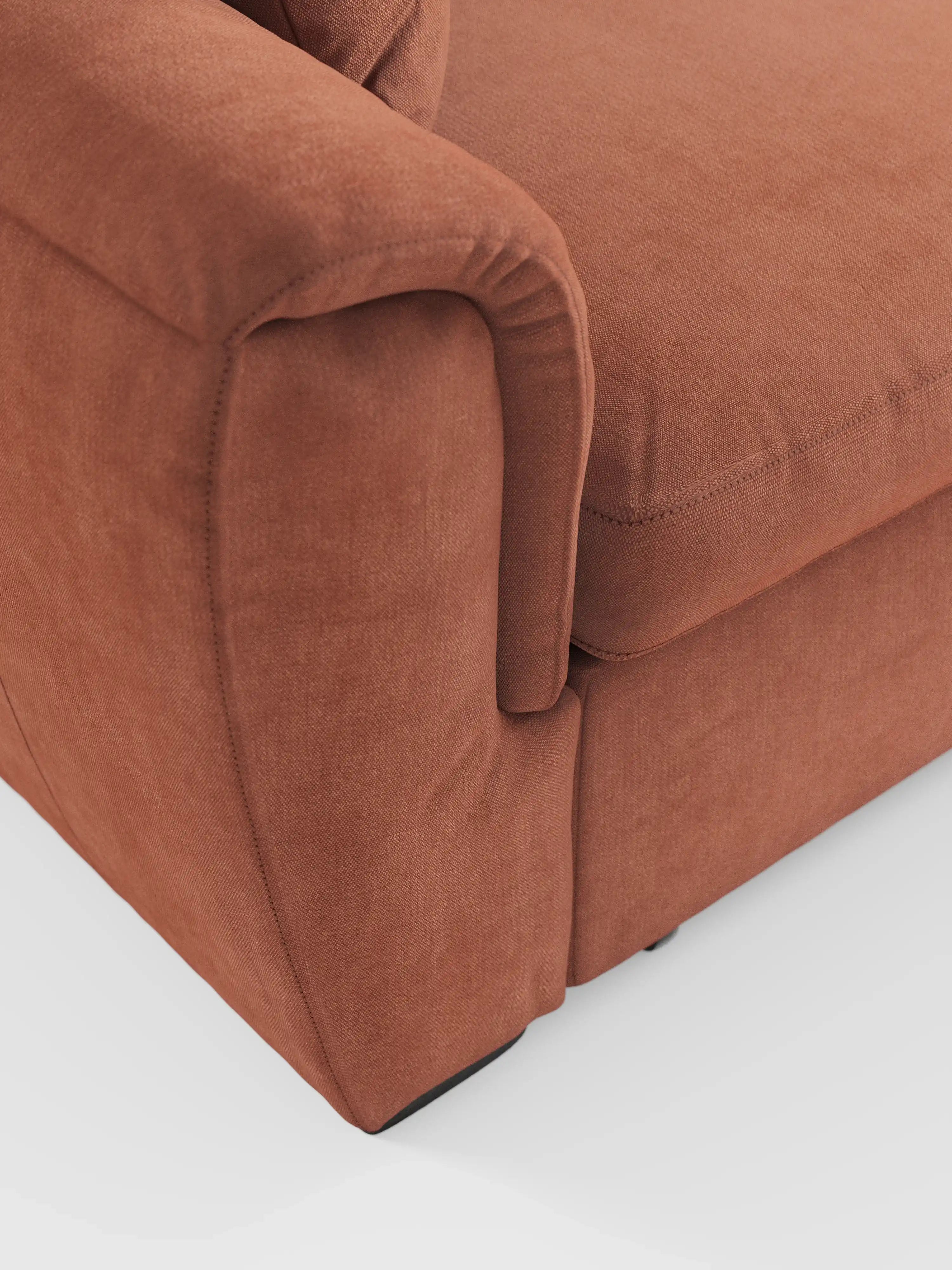 Primo piano dell'angolo anteriore del divano letto Titano con penisola reversibile e contenitore di Divani.Store, realizzato in tessuto color ruggine e resistente alle macchie, che mostra un bracciolo, le cuciture a vista e parte del cuscino della seduta. Lo sfondo è una superficie bianca liscia.