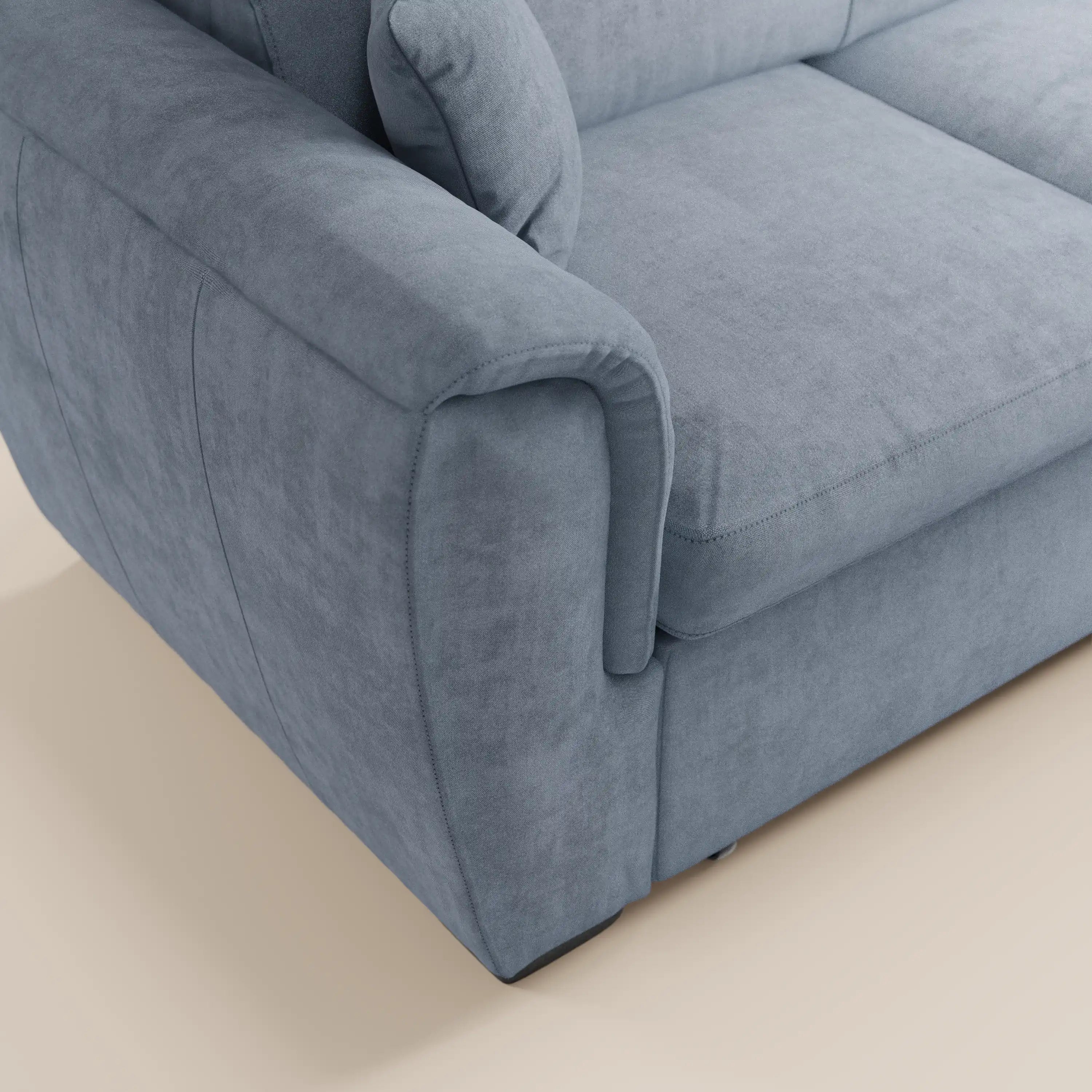 Primo piano del bracciolo e del cuscino di seduta del divano letto Titano con penisola reversibile, contenitore, poggiatesta reclinabili ed elemento estraibile in tessuto smacchiabile di Divani.Store, qui presentato con un rivestimento in peluche azzurro chiaro dalla consistenza morbida e vellutata. Il divano dal design moderno poggia su un pavimento beige chiaro e presenta cuciture a vista lungo i bordi che gli conferiscono un aspetto raffinato e sartoriale.