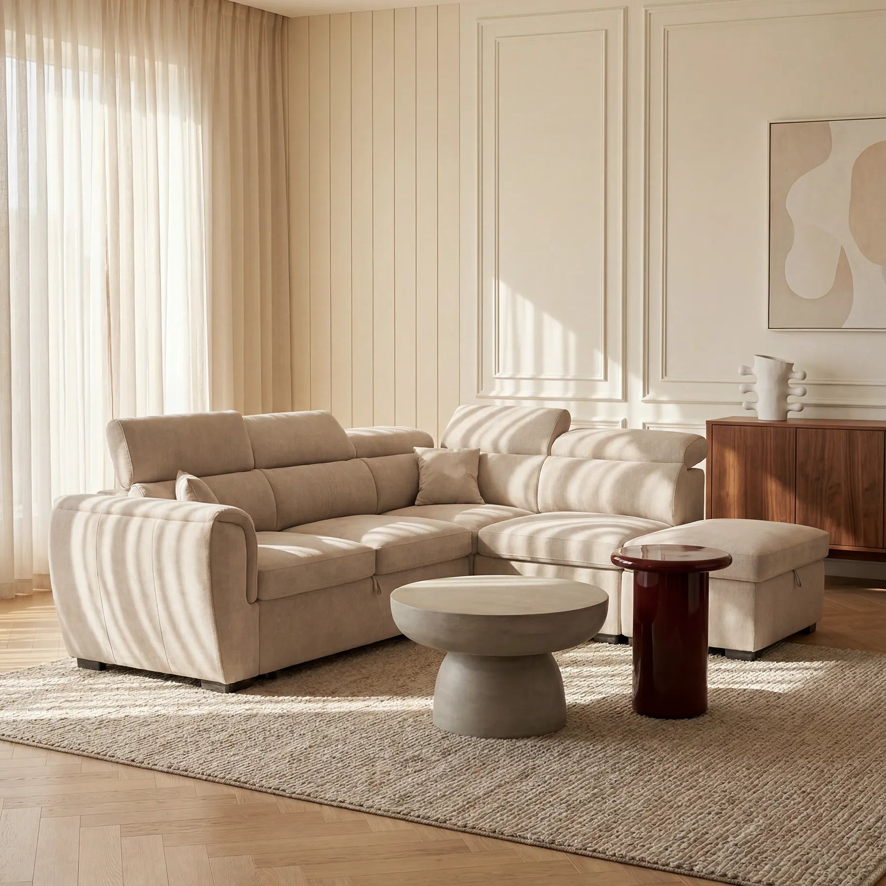 Un soggiorno moderno con il divano angolare reversibile Titano con letto estraibile e pouf contenitore di Divani.Store, tavolino rotondo in cemento, tavolino lucido e pouf su un tappeto intrecciato. La luce filtra attraverso le tende trasparenti; le pareti presentano modanature a pannelli e opere d’arte astratta.
